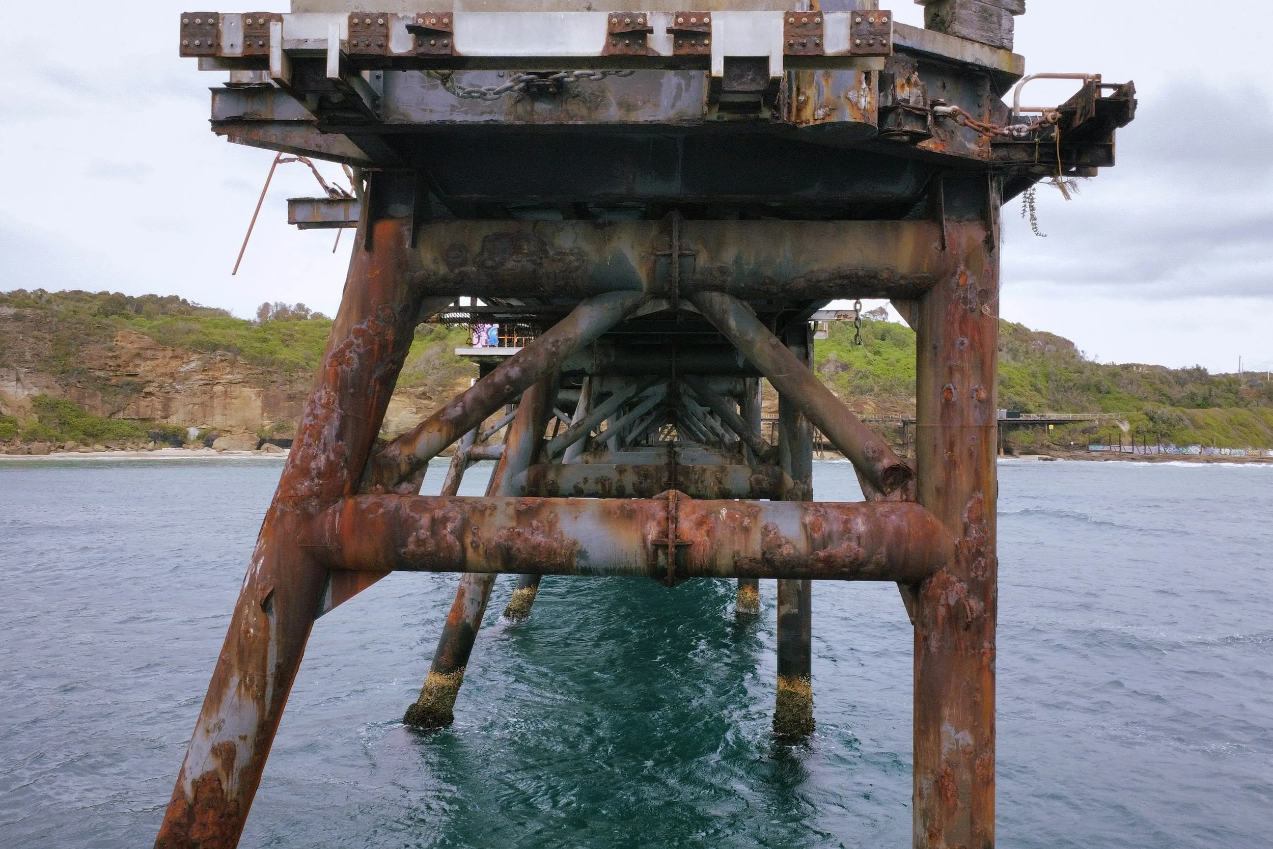Underwater view of a rusty metal pier extending into the water, with hills and cliffs in the background.
