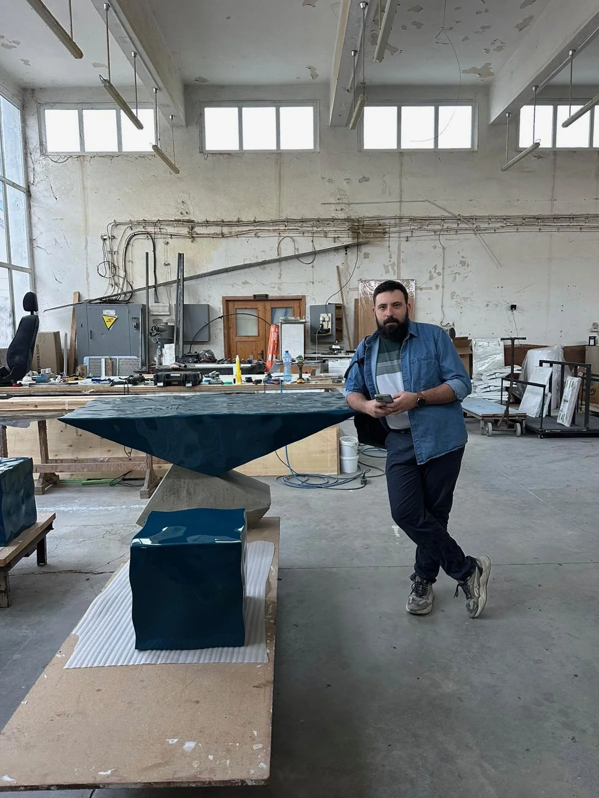 A man with a beard, wearing a blue denim jacket, striped shirt, black pants, and sneakers, is standing inside an industrial workshop with high ceilings, large windows, and exposed wires. He is leaning against a large table with a dark blue object on it, holding a cellphone in his right hand.