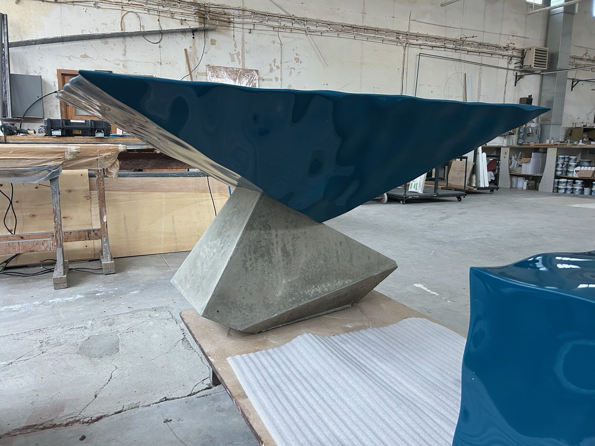 A modern sculpture consisting of a large blue, glossy, wave-like structure mounted on a geometric concrete base in an industrial workshop setting.