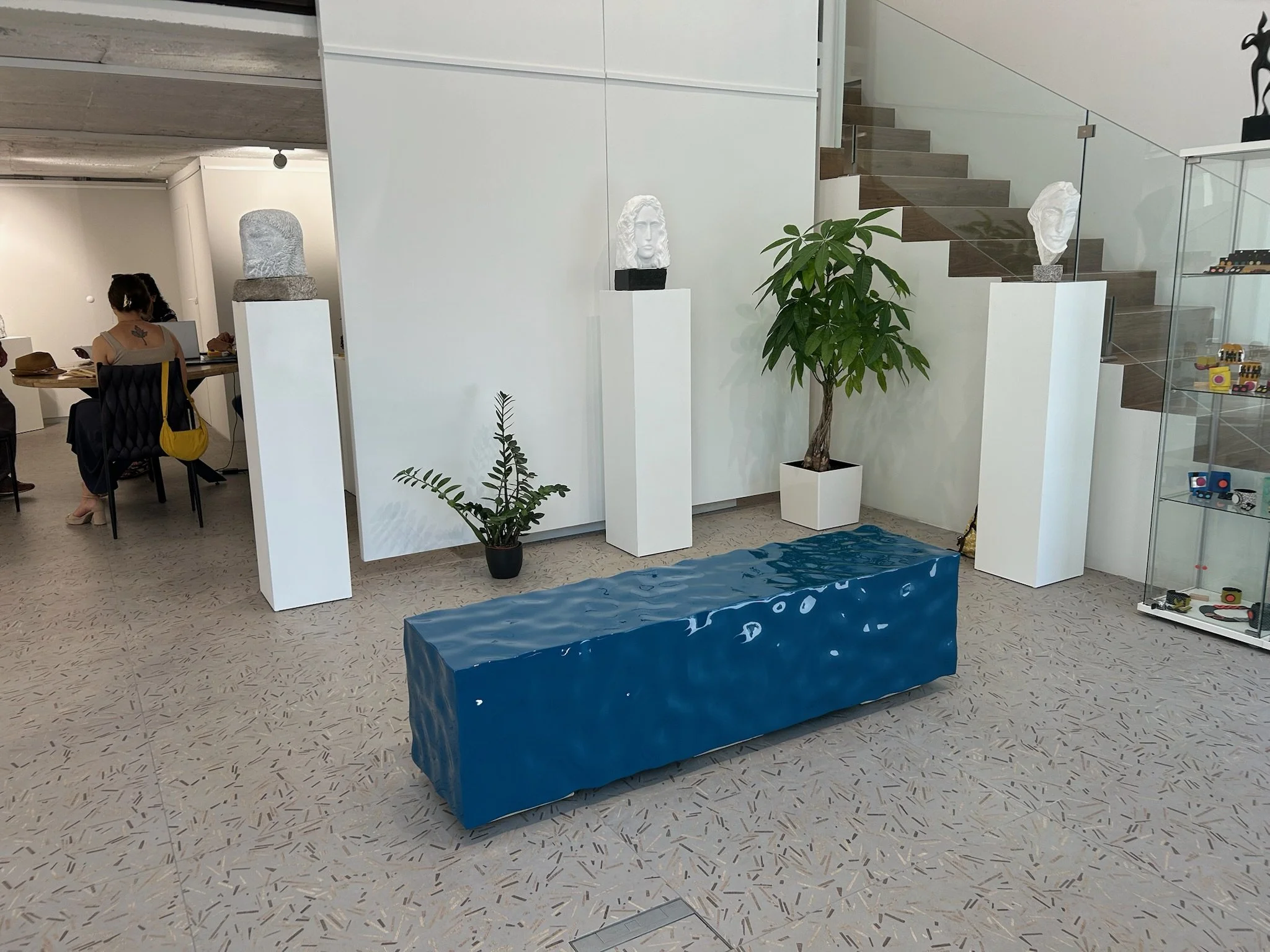 Indoor space with a blue textured bench, two white pedestals with white bust sculptures, a potted plant, and a staircase with glass railing.