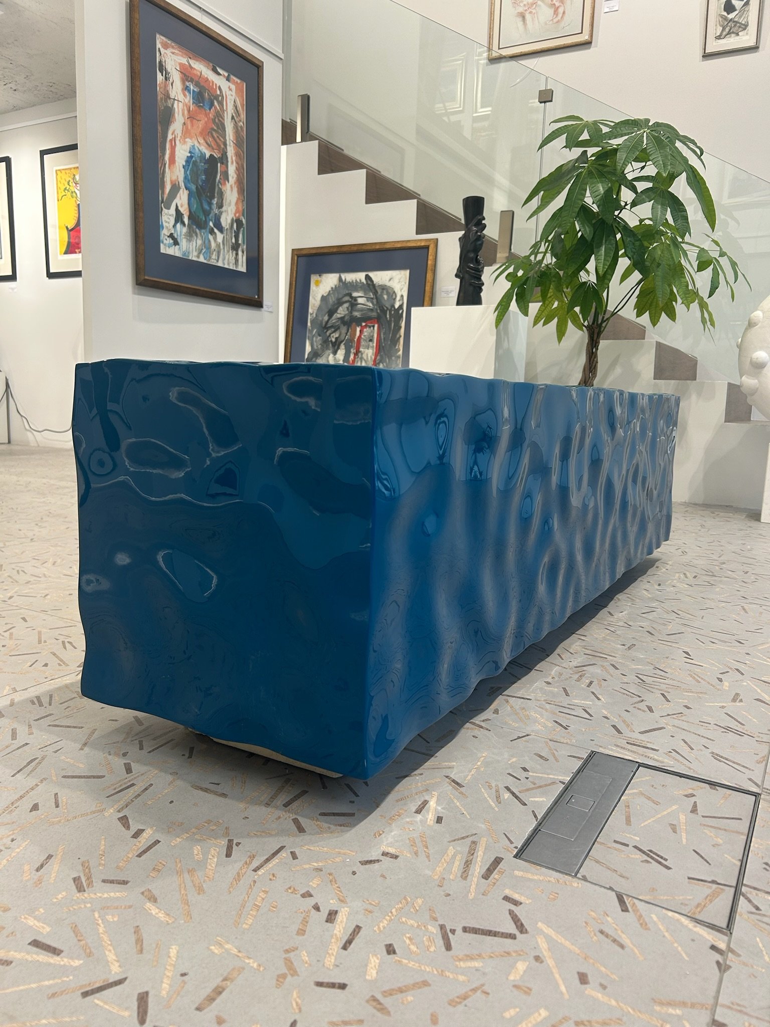 Modern art gallery interior featuring a large blue, textured bench, framed abstract paintings on the wall, a potted plant next to a staircase, and a terrazzo floor. In a gallery.