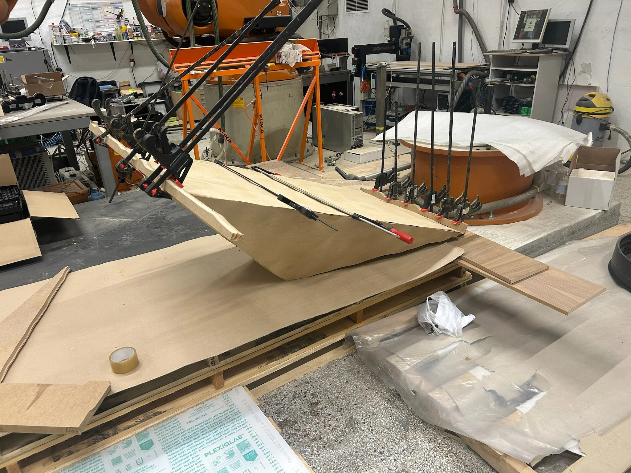 Workshop or laboratory with a wooden boat under construction, clamps holding the boat's parts, various tools, materials, and equipment scattered around.