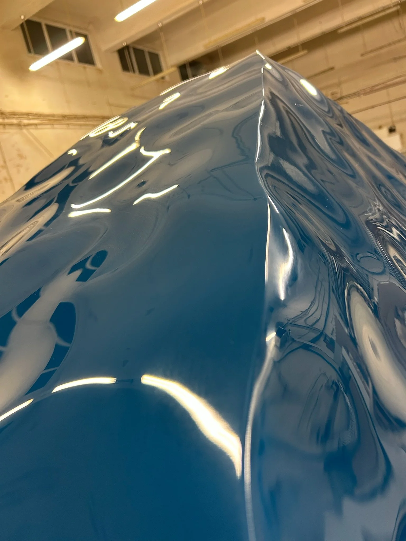 Close-up of a shiny, reflective, metallic blue sculpture or object with smooth curves in an indoor space, with ceiling lights and windows in the background.