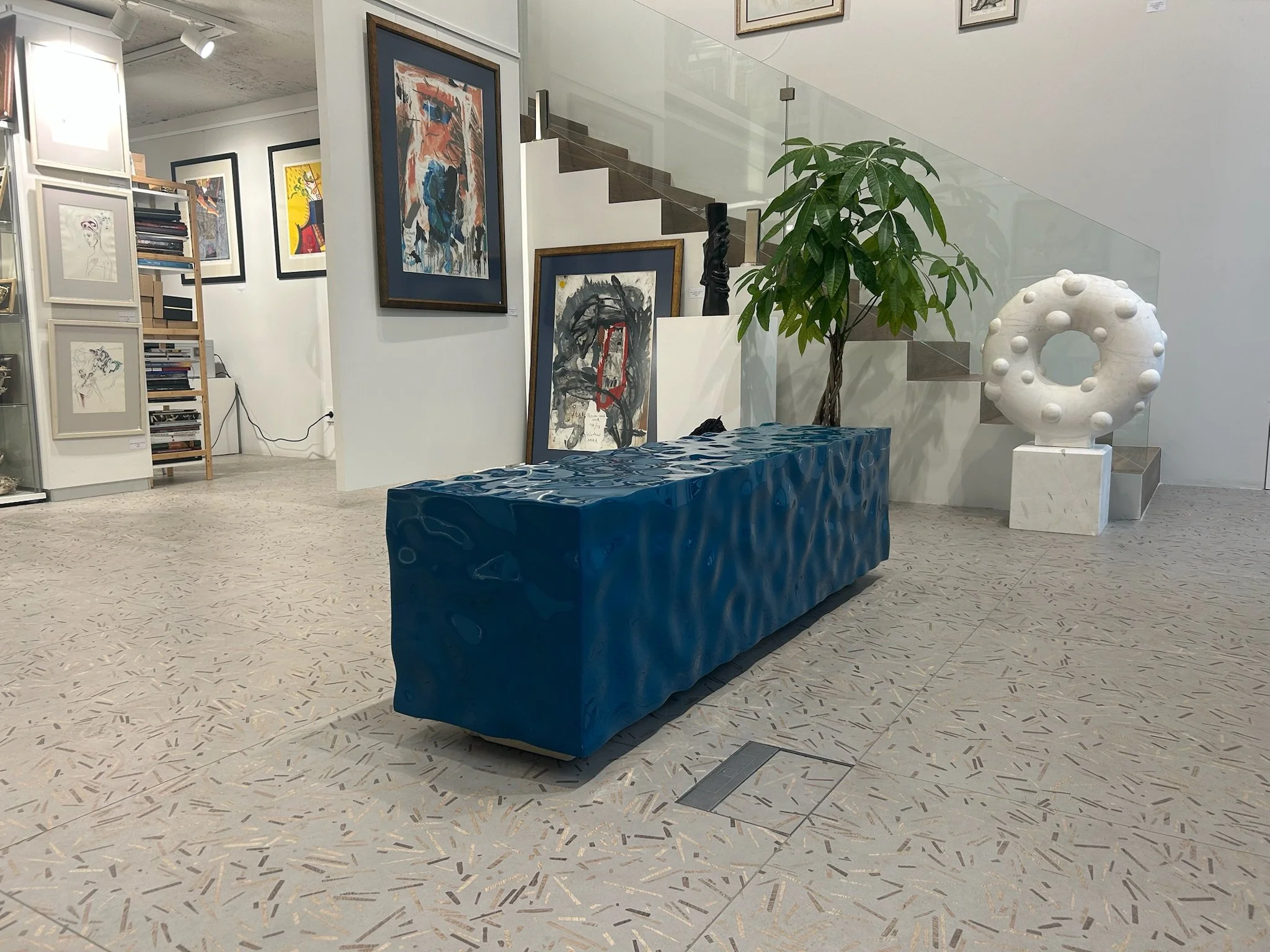 Interior view of an art gallery showing abstract paintings on the walls, a blue textured bench, a potted plant, and a white sculpture of a donut shape with round protrusions.