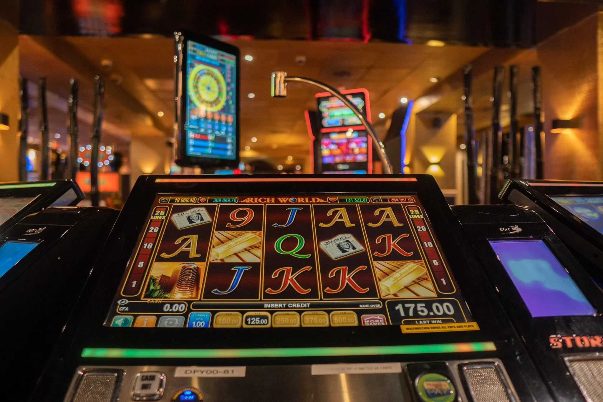 Slot machine displaying a game with multiple reels showing cards and symbols, in a casino with other gaming machines and colorful lights in the background.