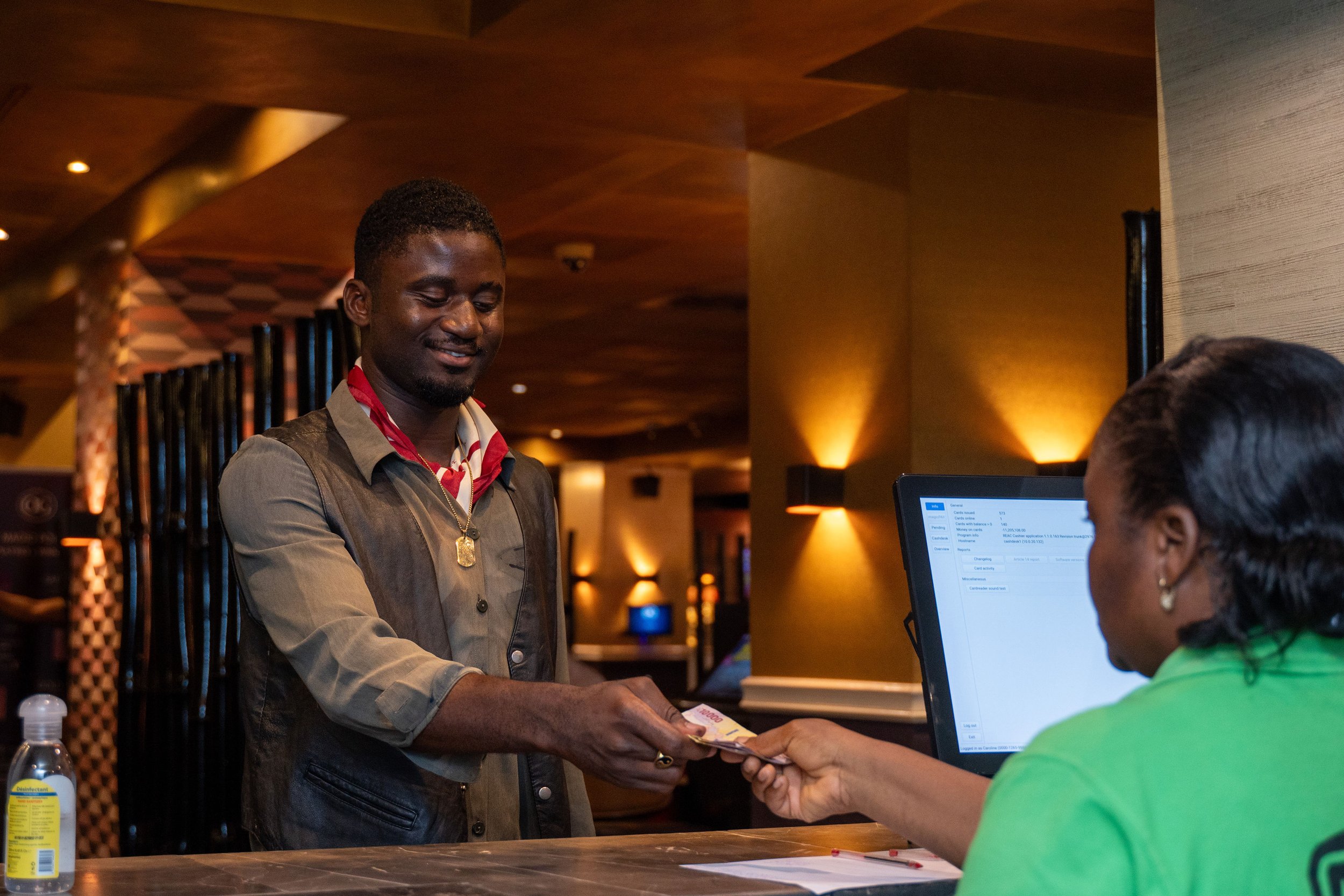 A man smiling and handing money to a woman at a reception desk in a warmly lit hotel lobby.