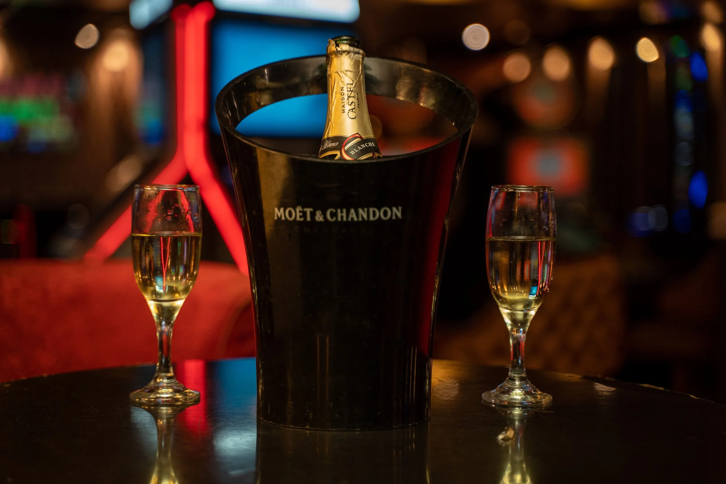 A bucket with a bottle of Moët & Chandon champagne inside, flanked by two glasses of champagne on a dark wooden table in a dimly lit bar or lounge with colorful blurred lights in the background.