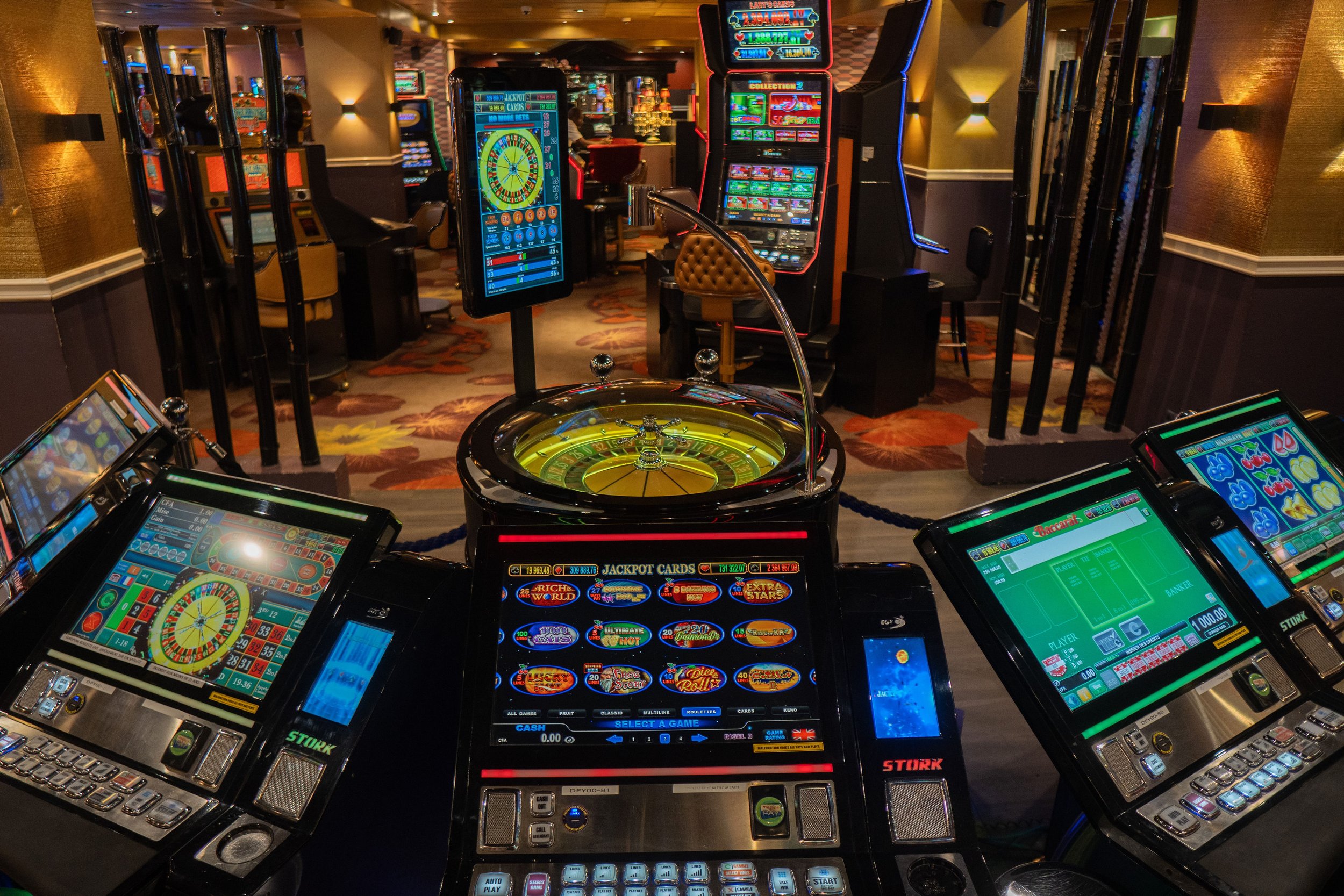 A row of slot machines and a roulette game in a casino with bright lights and colorful screens.