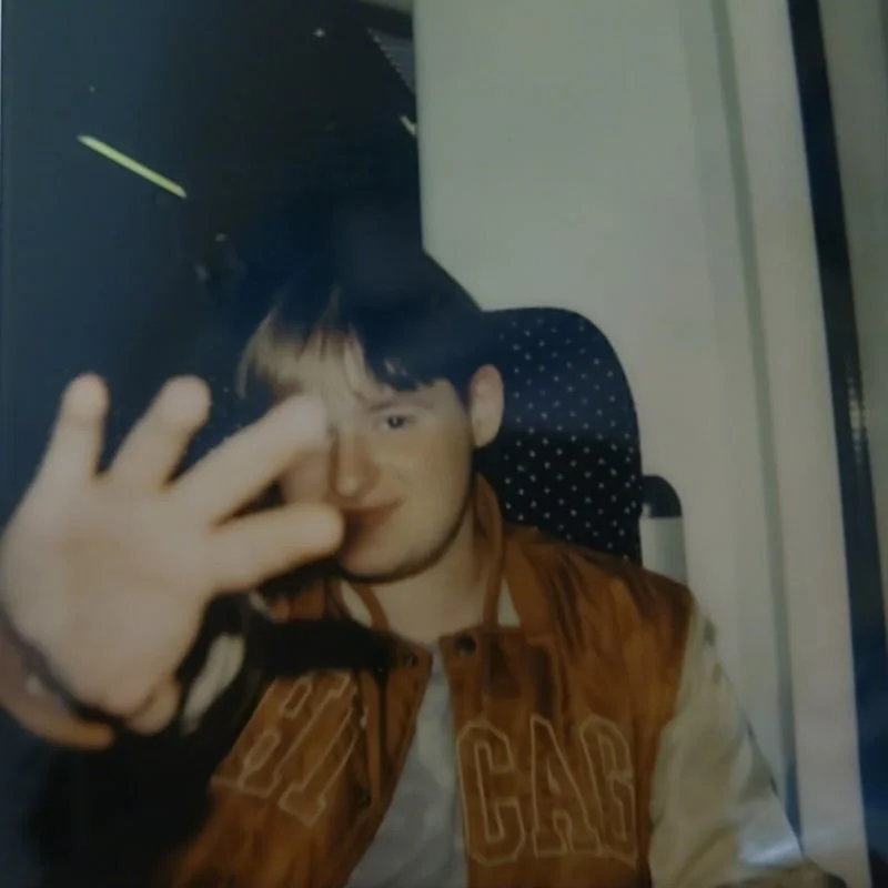 Junge Person sitzt in einem Zug, trägt eine braune College-Jacke und blickt lächelnd in die Kamera, während er die Hand hochhält.