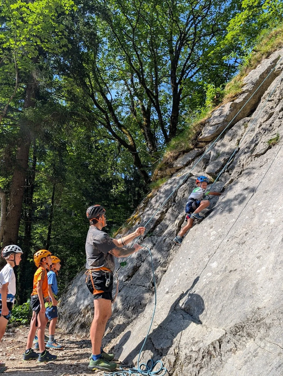 Climbing harnesses and gear laid out on the ground in a forested outdoor setting, with four people standing in the background.