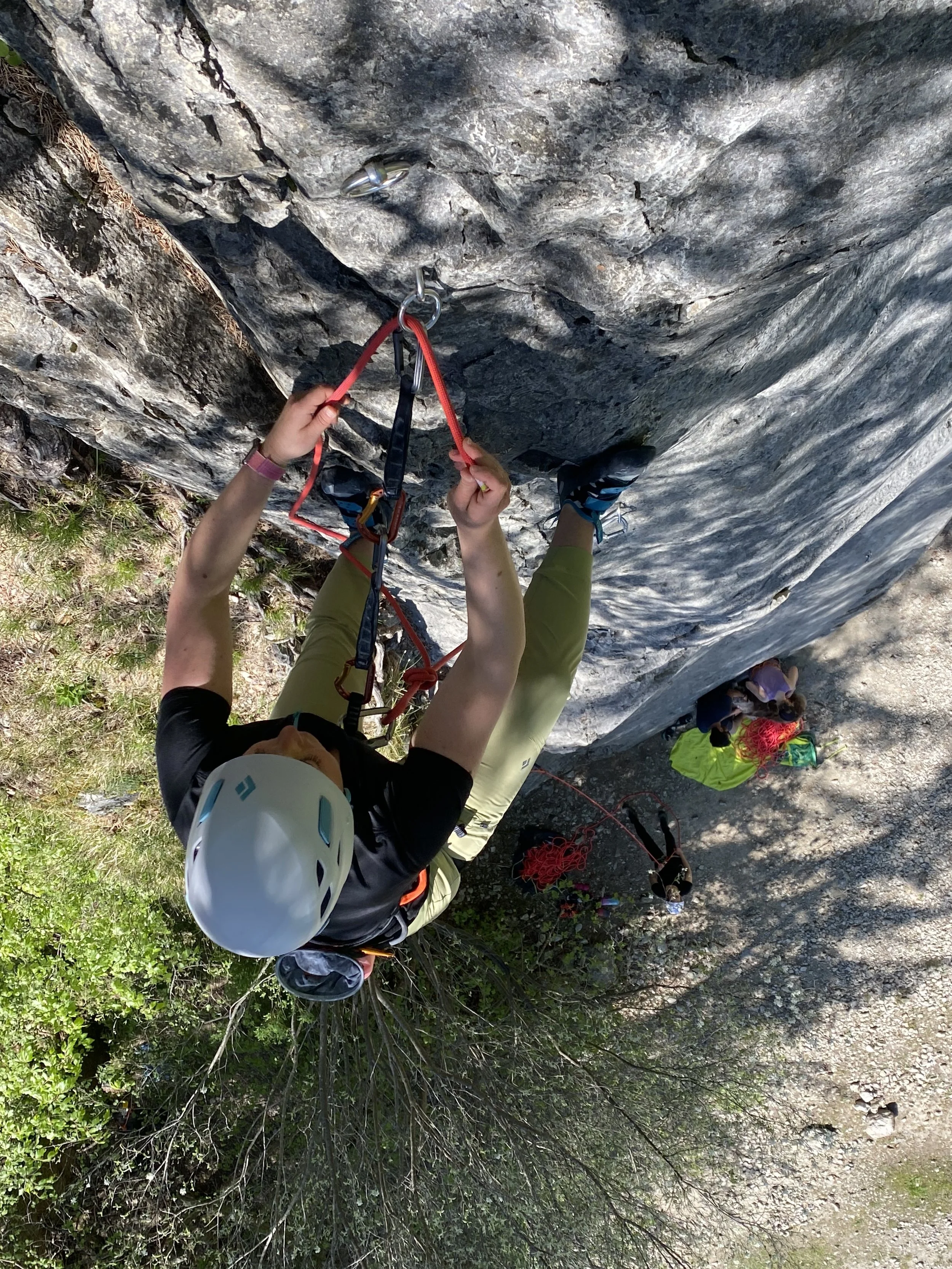 A person rock climbing on a steep rock face wearing a helmet and harness, with climbing gear, while others wait below. The view is from above showing the climber's perspective.