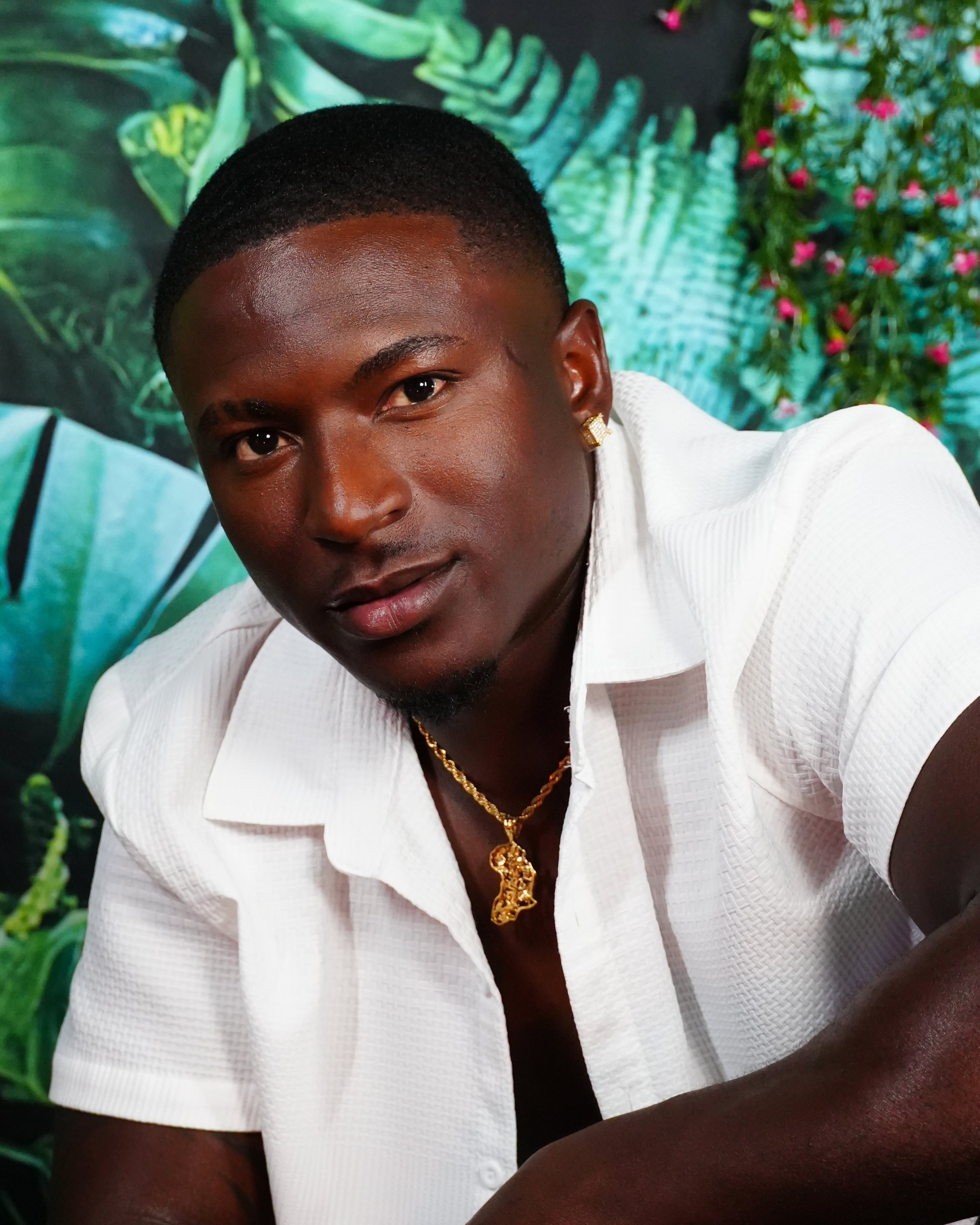 A young man with dark skin, short haircut, and wearing a white shirt, gold jewelry, and earrings, posing in front of a lush green jungle background.