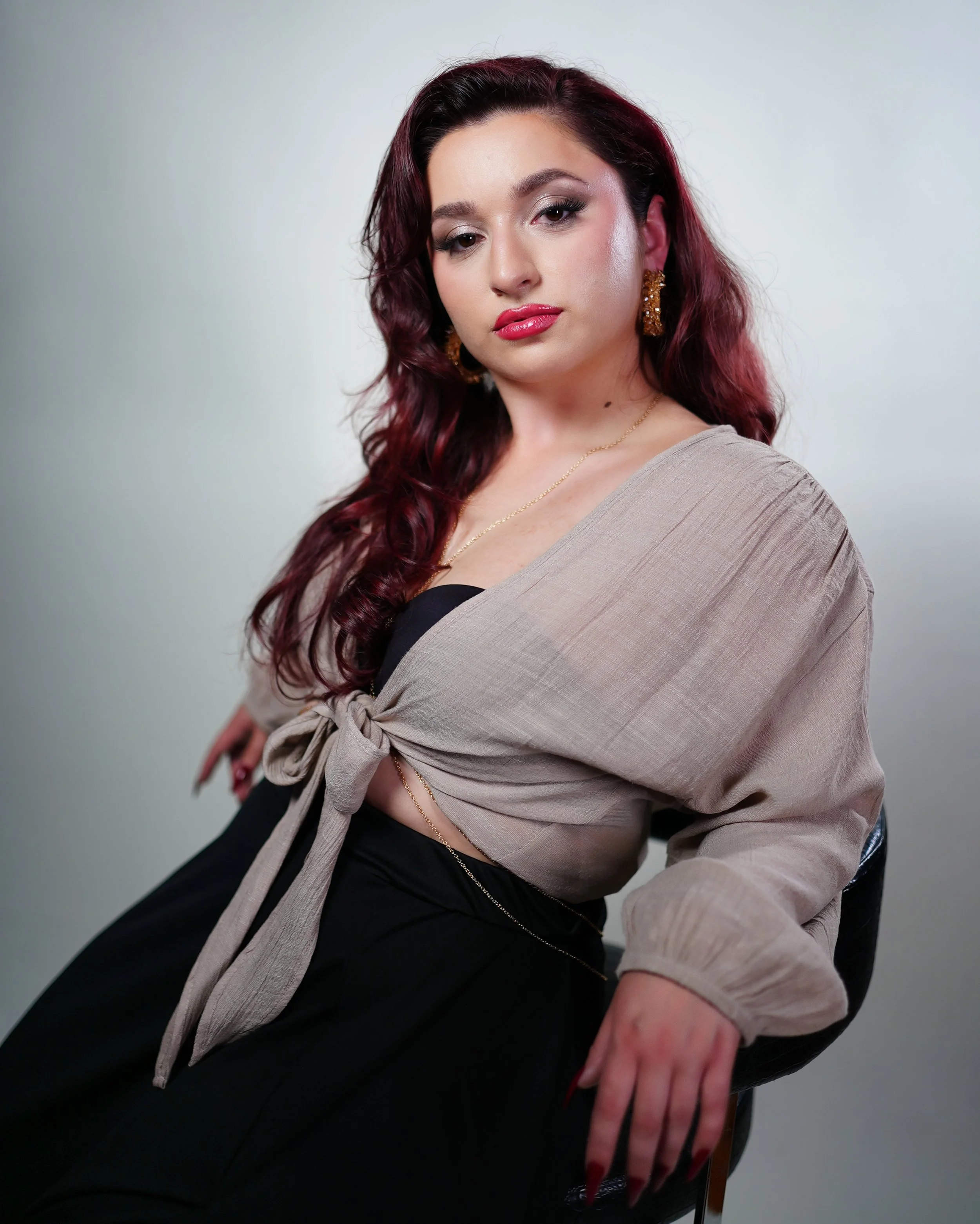 A young woman with long, curly, dark red hair and makeup, wearing a black top, a beige tied-up shirt, and gold jewelry, seated in a black chair against a plain light gray background.