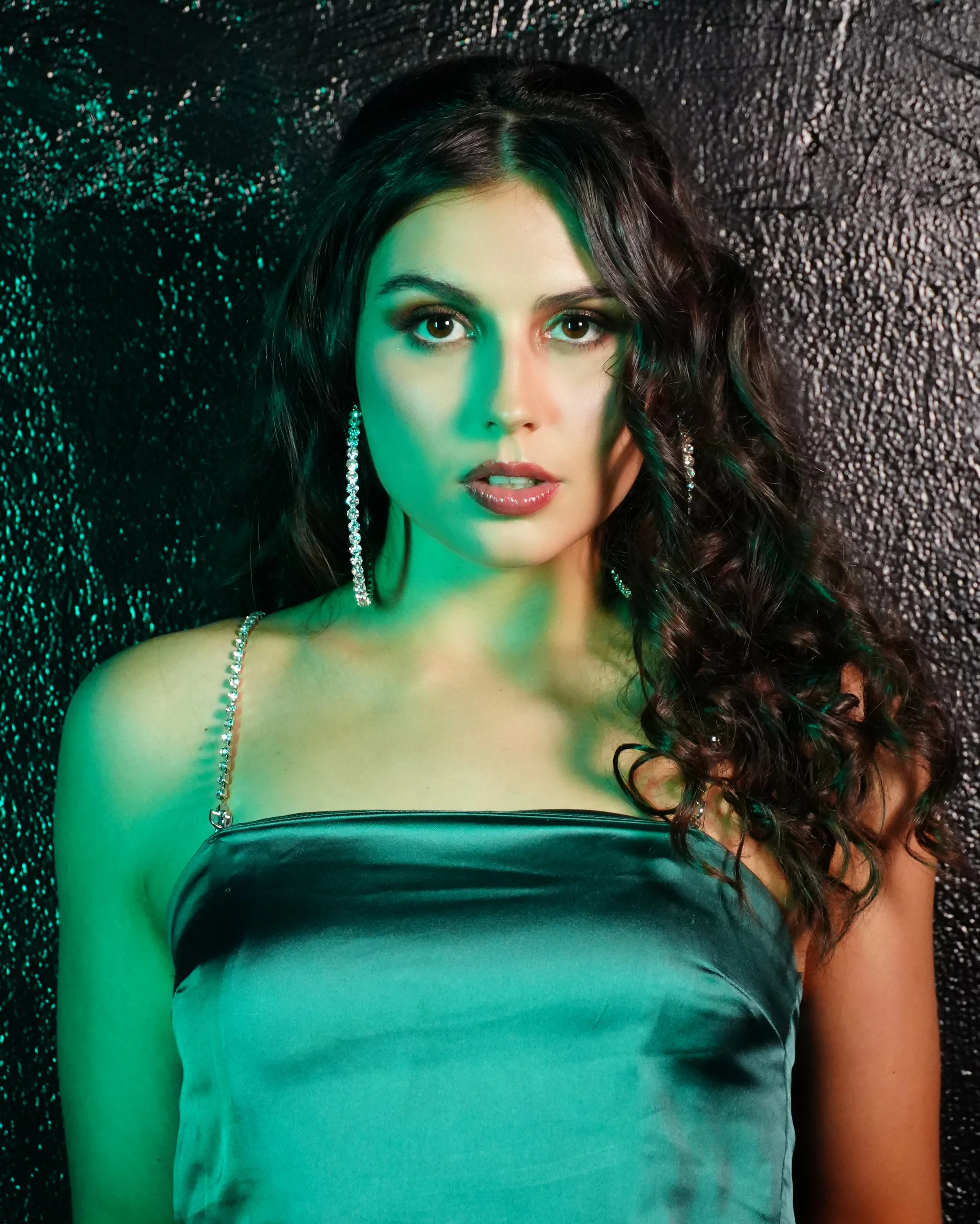 Portrait of a woman with dark, curly hair, wearing a strapless satin dress and long, dangling earrings, standing against a dark, textured background.