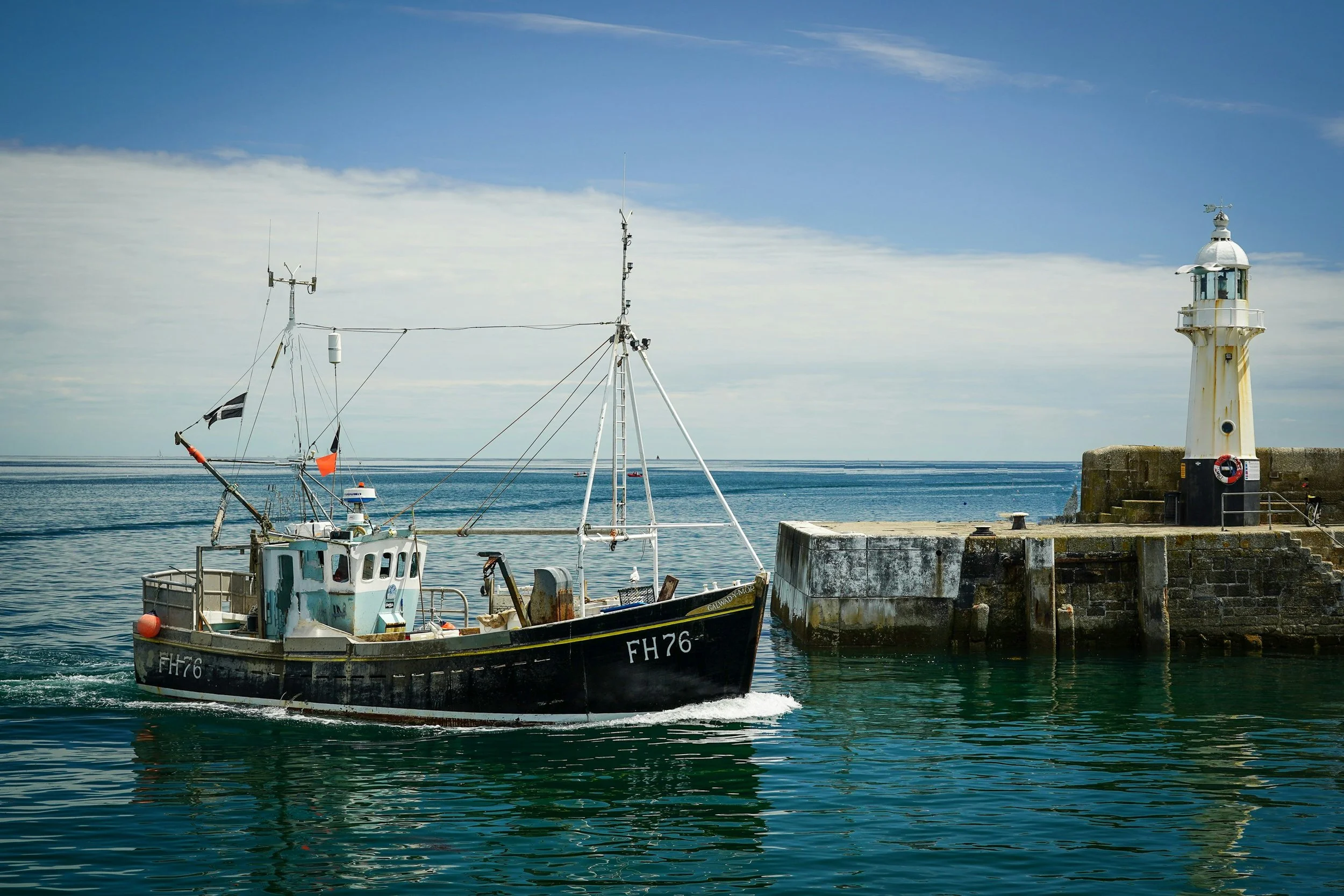 A small fishing boat with the code FH76 docked near a lighthouse on a harbor, with a calm sea and partly cloudy sky in the background.
