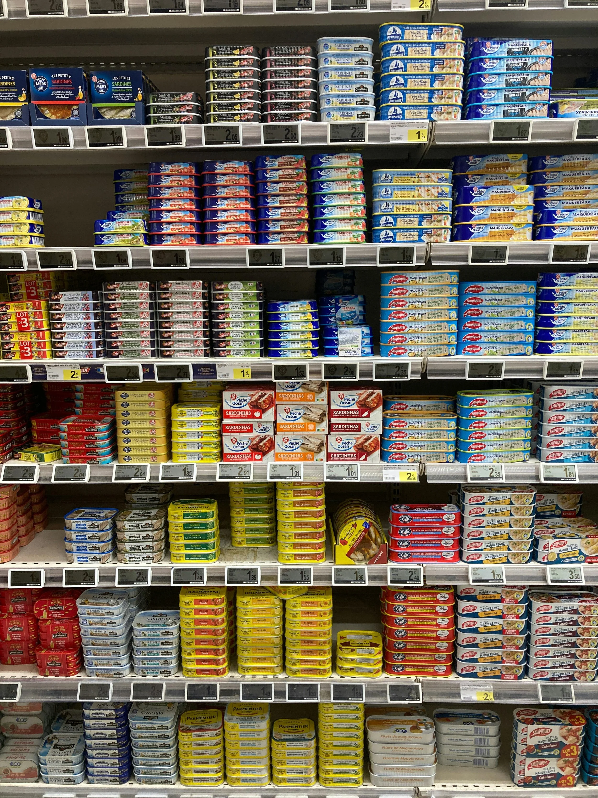 Shelf in a grocery store with various canned seafood products, mainly sardines, stacked in colorful tins.