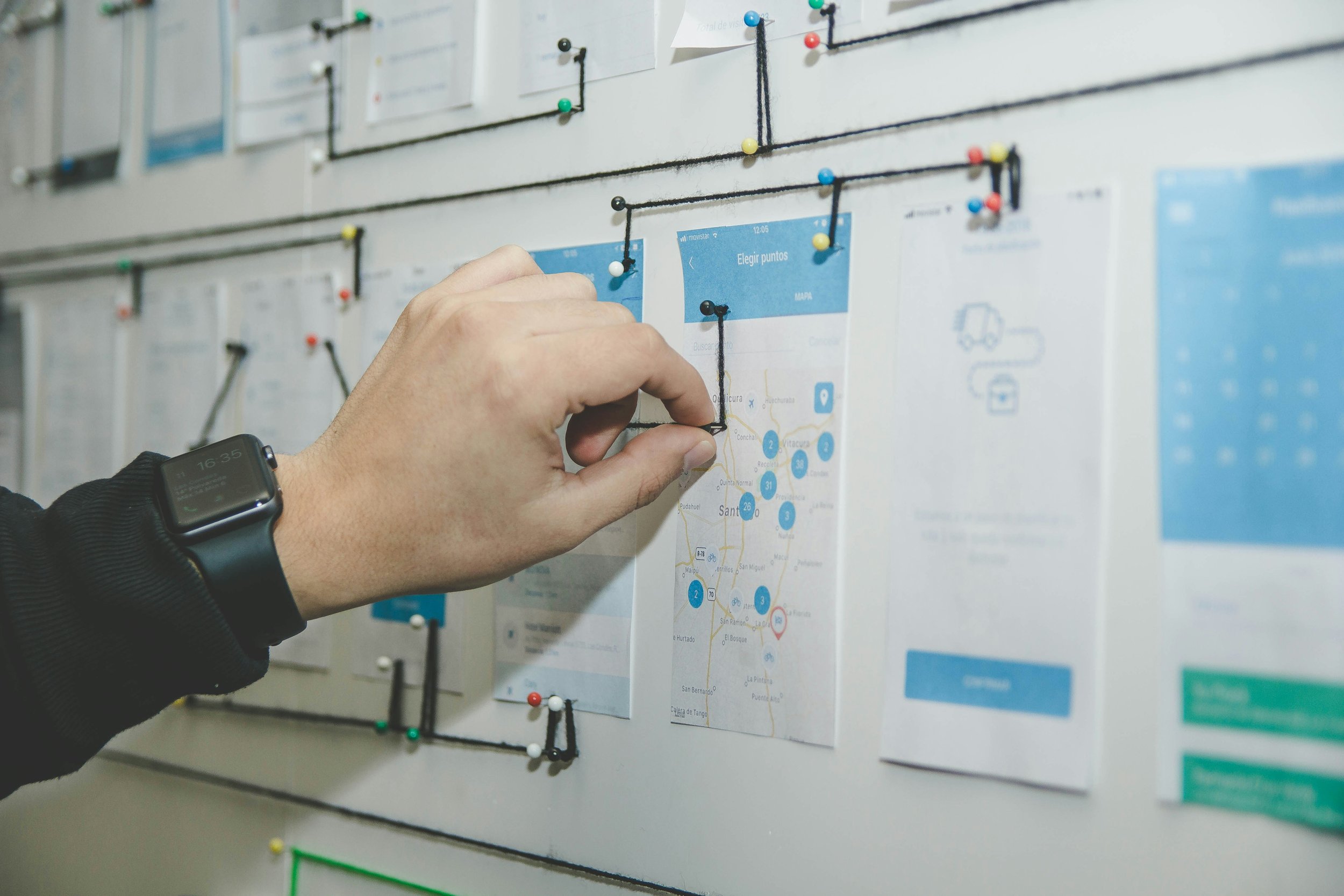 A person's hand is pinning a paper map to a bulletin board using push pins. The person is wearing a black smartwatch. The bulletin board has several documents attached with push pins and black drawstrings, and some of the documents include maps and diagrams.