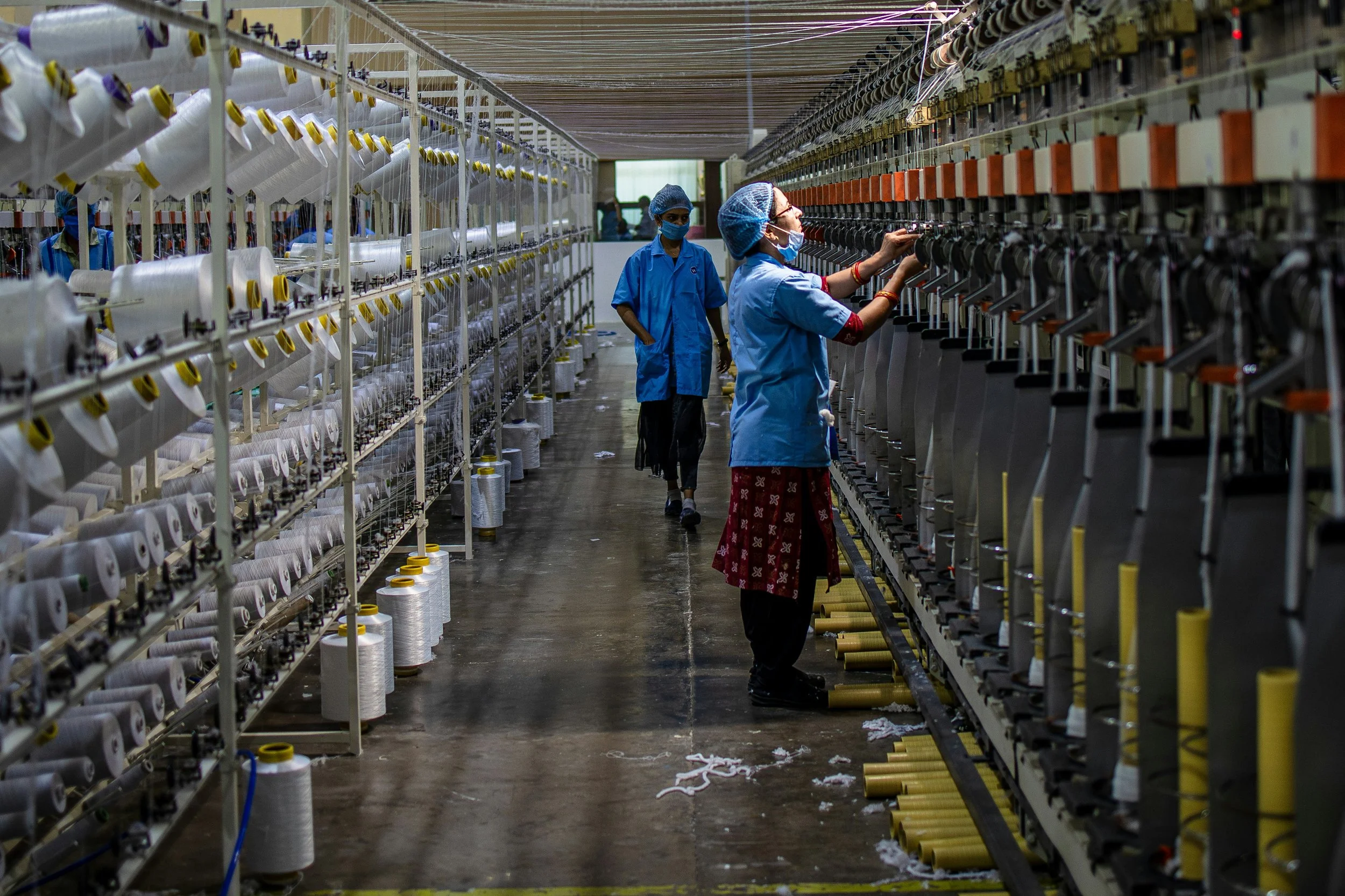 Workers in blue uniforms and face masks operate and inspect textile machines in a factory setting with spools of thread on shelves.