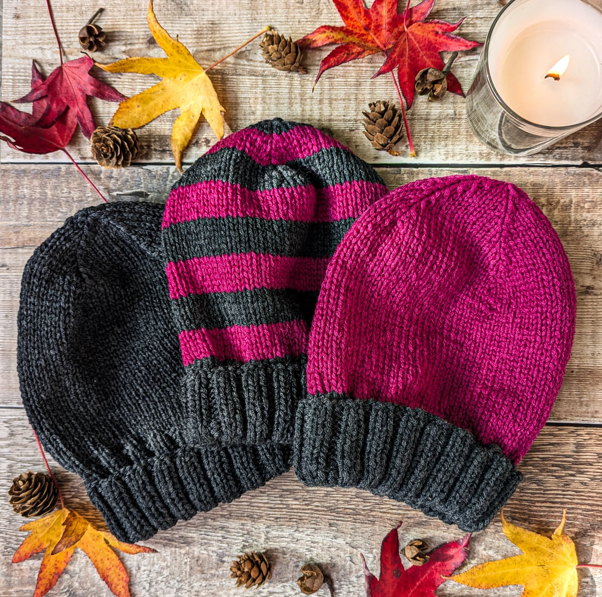 3 grey and pink knitted beanies on a wooden surface with leaves and a candle around them