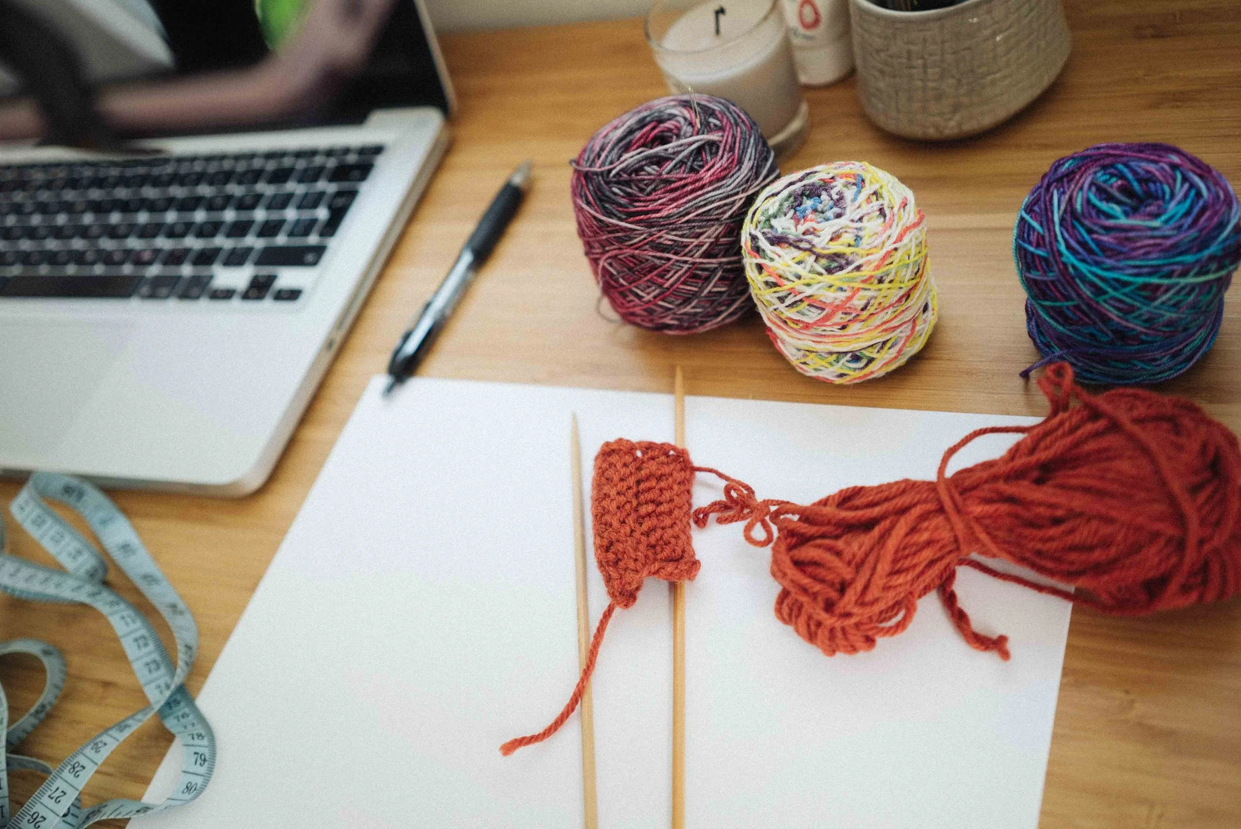 Colorful balls of yarn, some knitted samples, knitting needles, measuring tape, and a laptop on a wooden table.