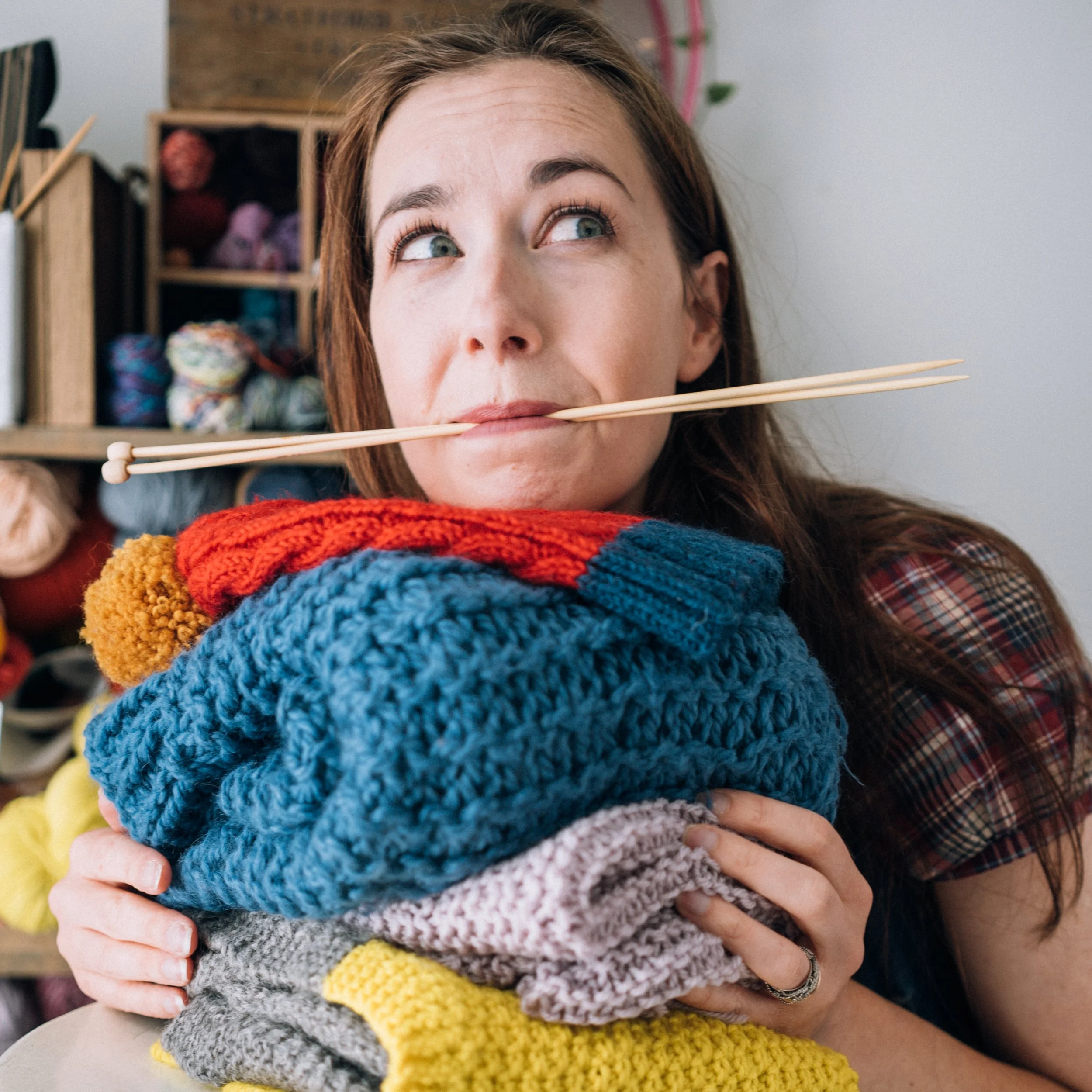Knitting Teacher Alice is partially behind a pile of handknitted items with wooden knitting needles in her mouth