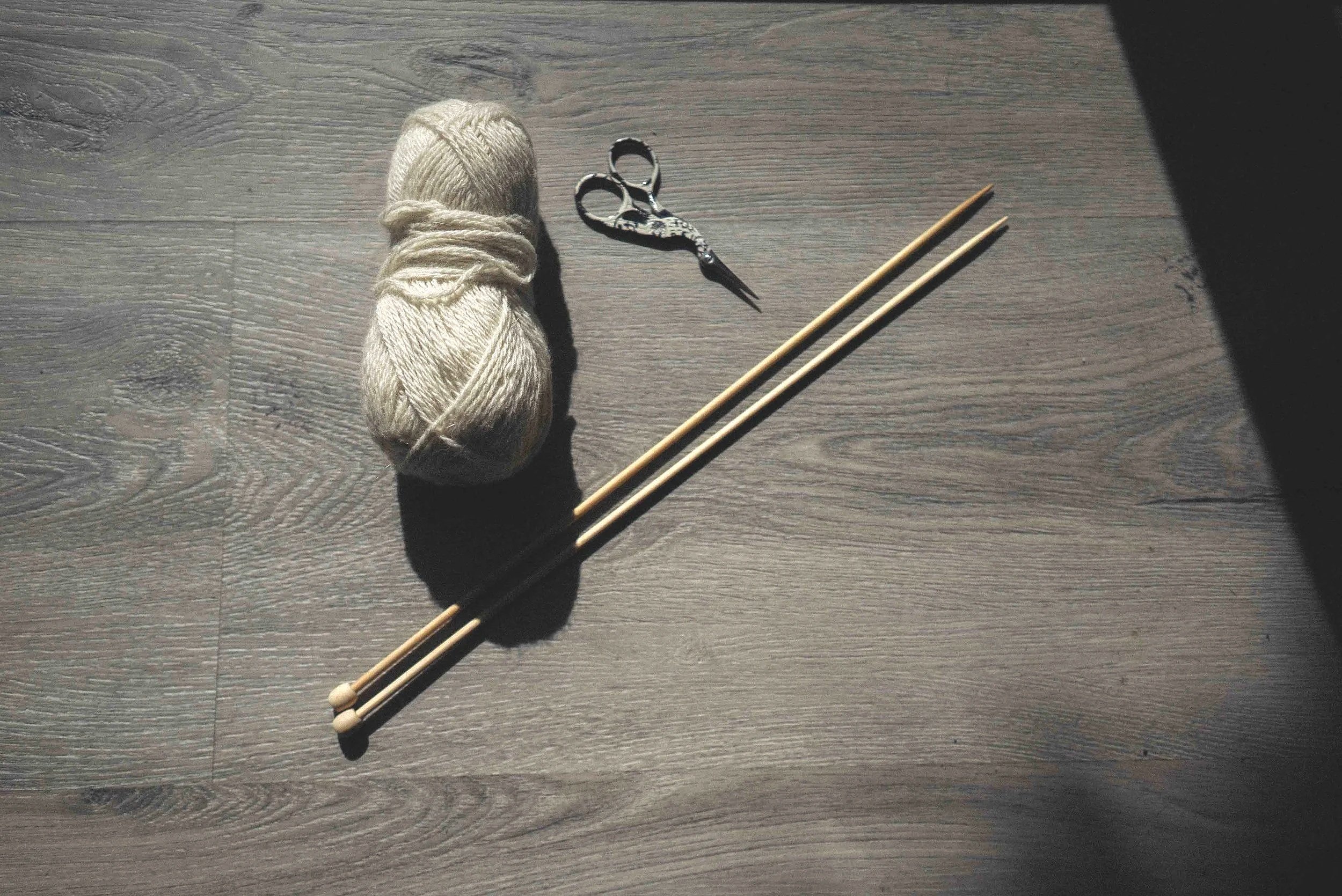A ball of beige yarn, a pair of black and silver scissors, and two light-colored knitting needles on a wooden surface.