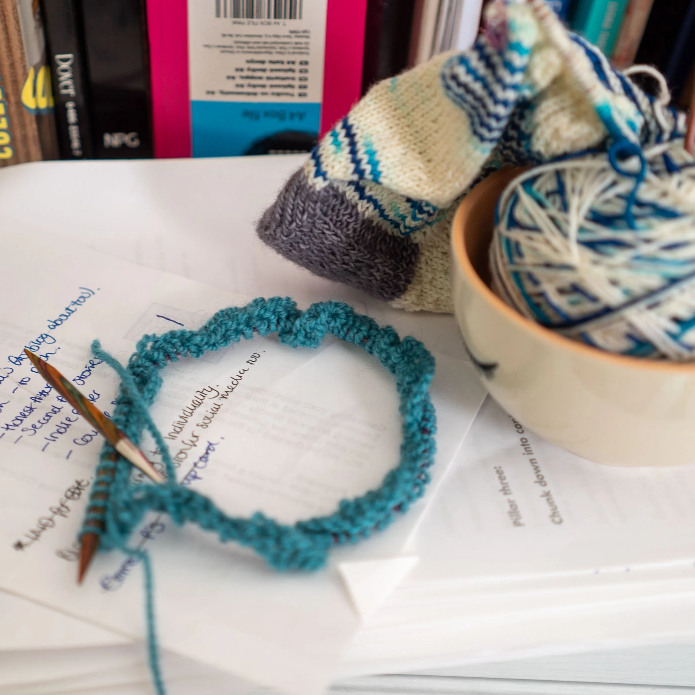 Close-up of a circular knitting project on a desk, with yarn and knitting needles, next to a notebook and books on shelves.