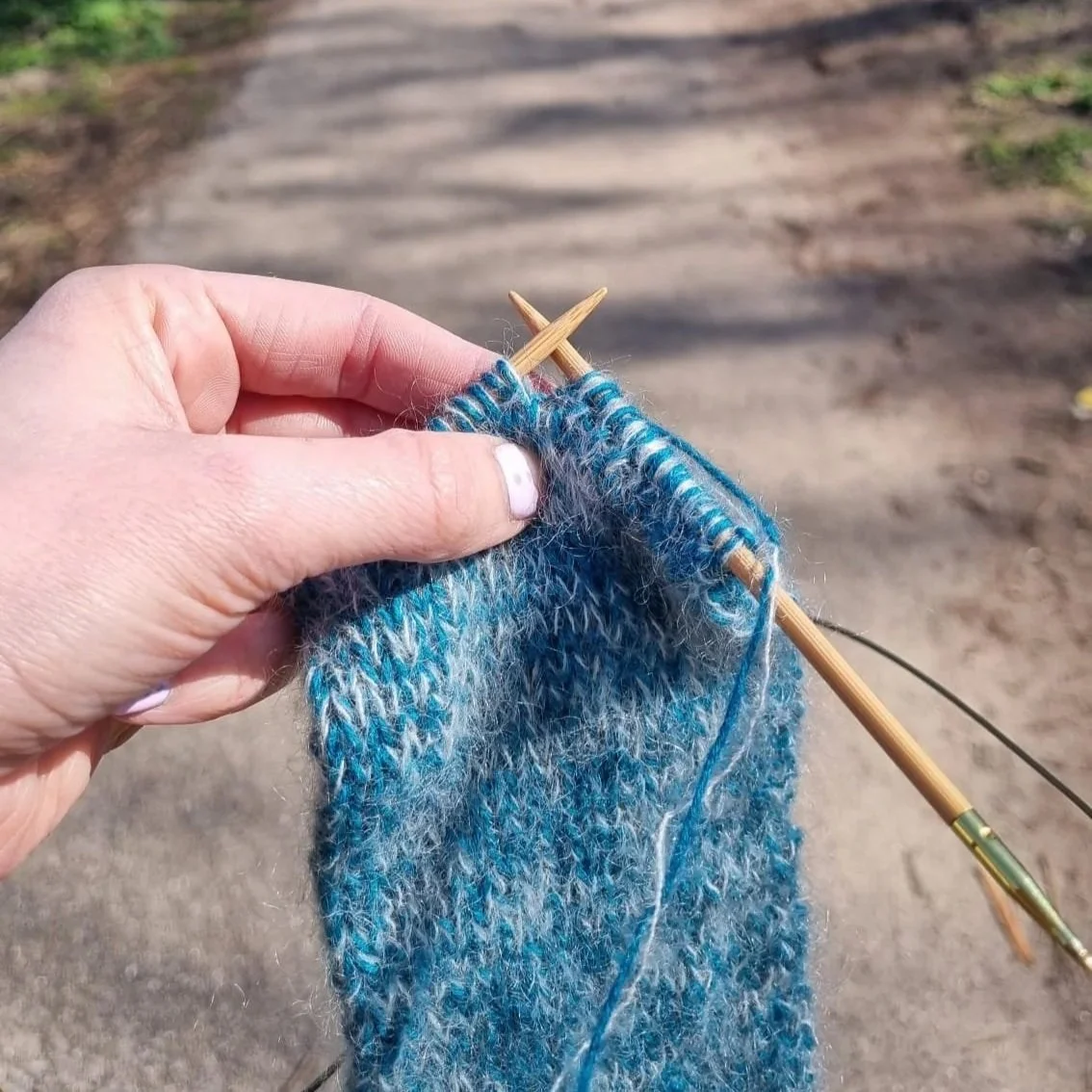 A white hand holds blue knitting on needles outside on a path