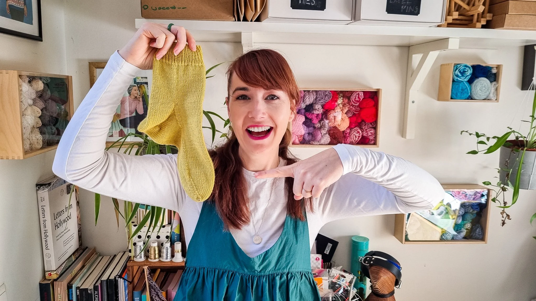 A woman with red hair smiling and pointing to a yellow knitted sock she is holding up, inside a yarn and craft store.