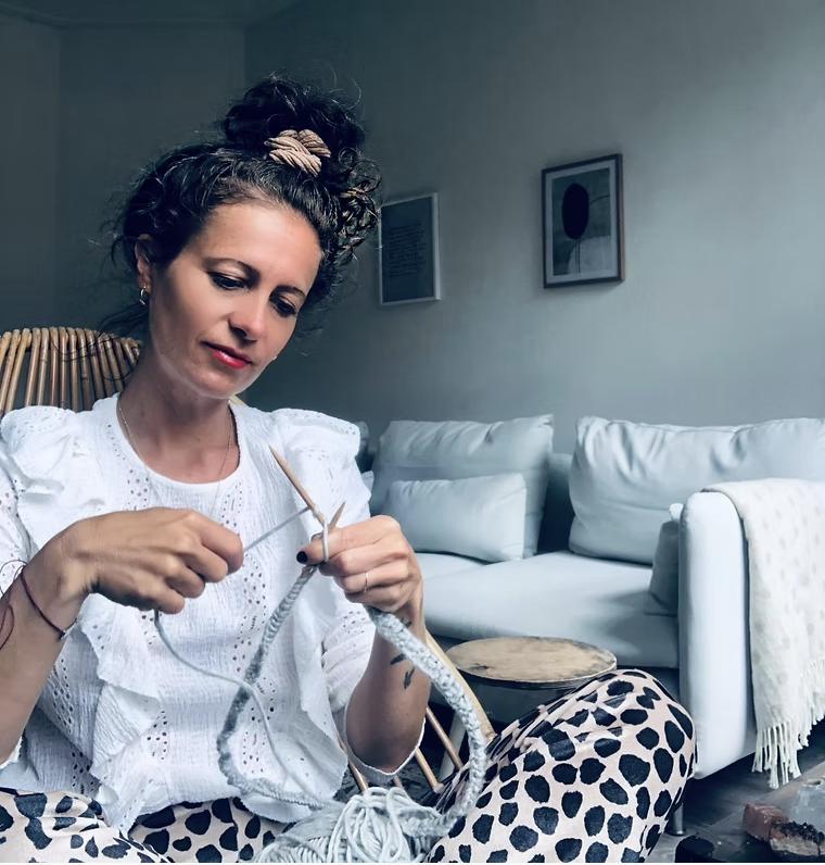 Claudia, a white female knits with a pale blue sofa behind her.