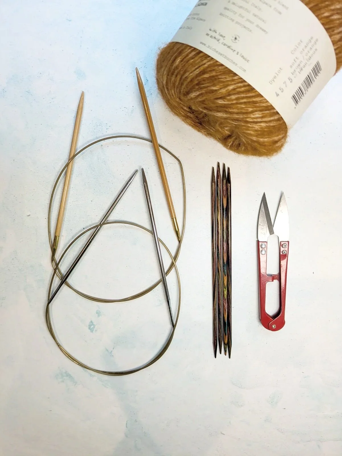 Set of knitting tools including circular knitting needles, double-pointed needles, a crochet hook, and a small pair of scissors, with a ball of brown yarn.