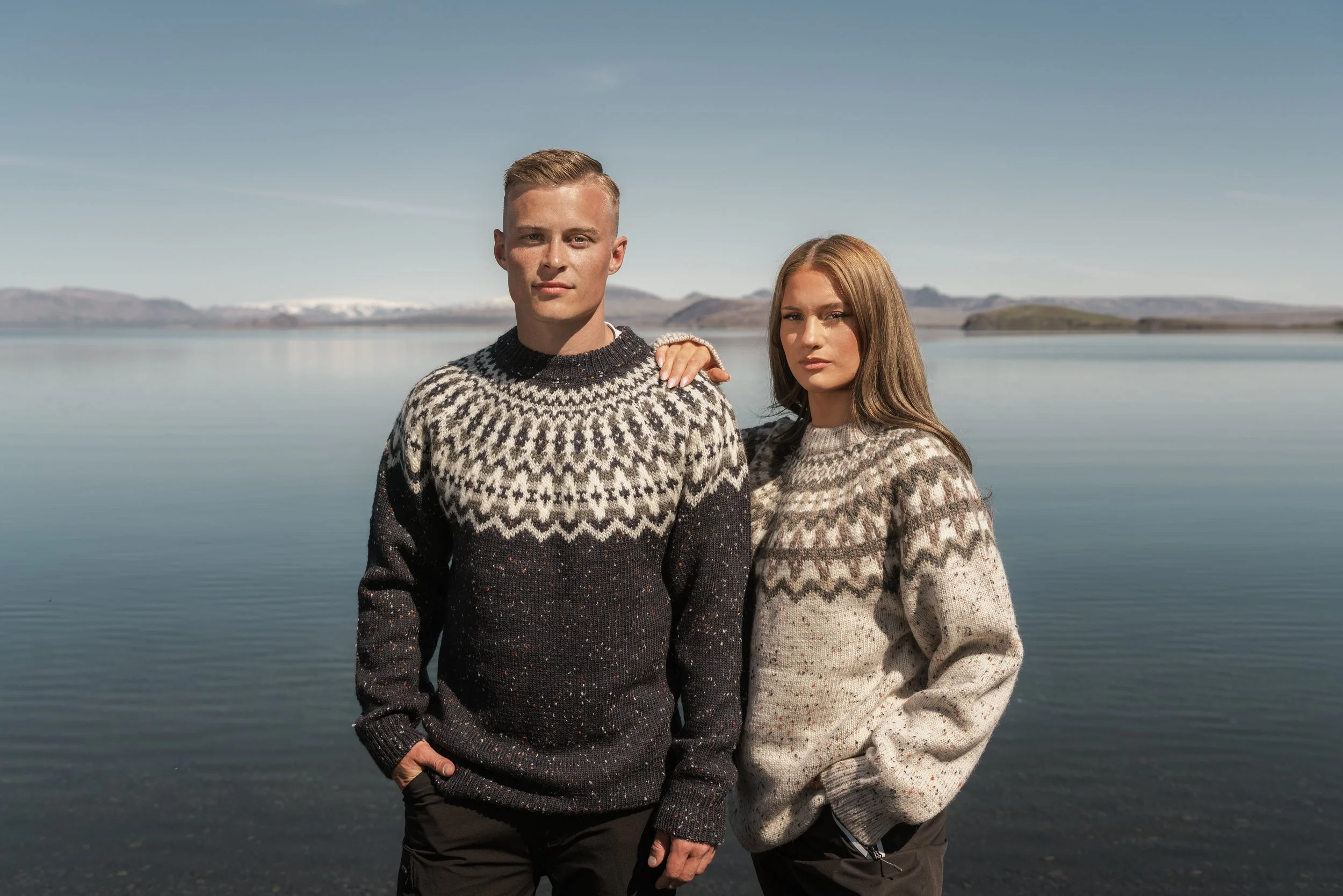 An Icelandic man and woman in classic icelandic knit sweaters stand in front of a large lake together