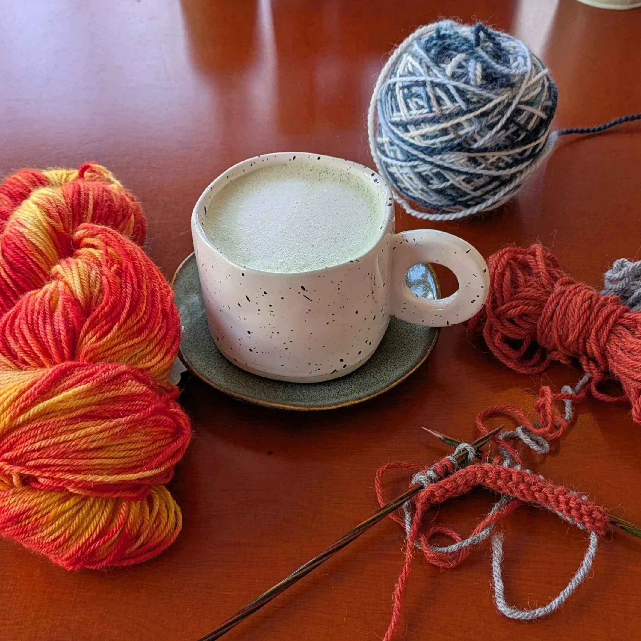 Balls of yarn in orange, gray, and multicolored on a wooden surface with knitting needles and a coffee mug.