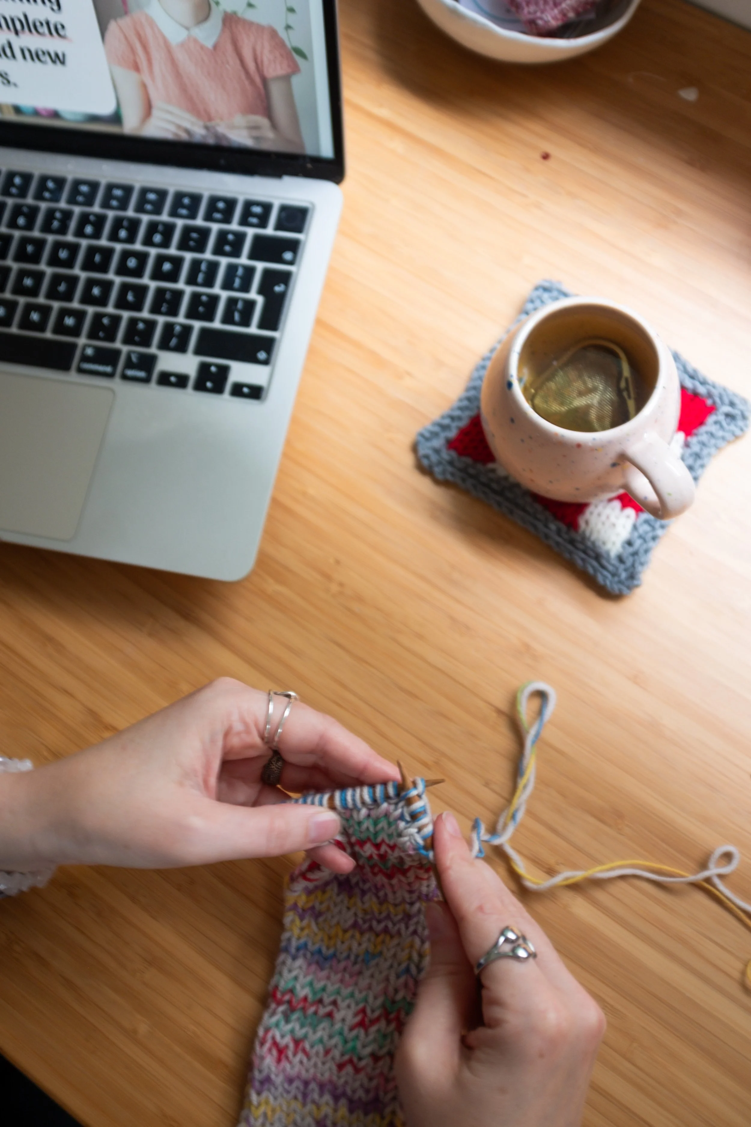 A woman knitting with multicolored yarn at a wooden desk. On the desk are a MacBook laptop, a blue measuring tape, and balls of yarn. The background shows a white wall with pink and white notes and papers pinned up.