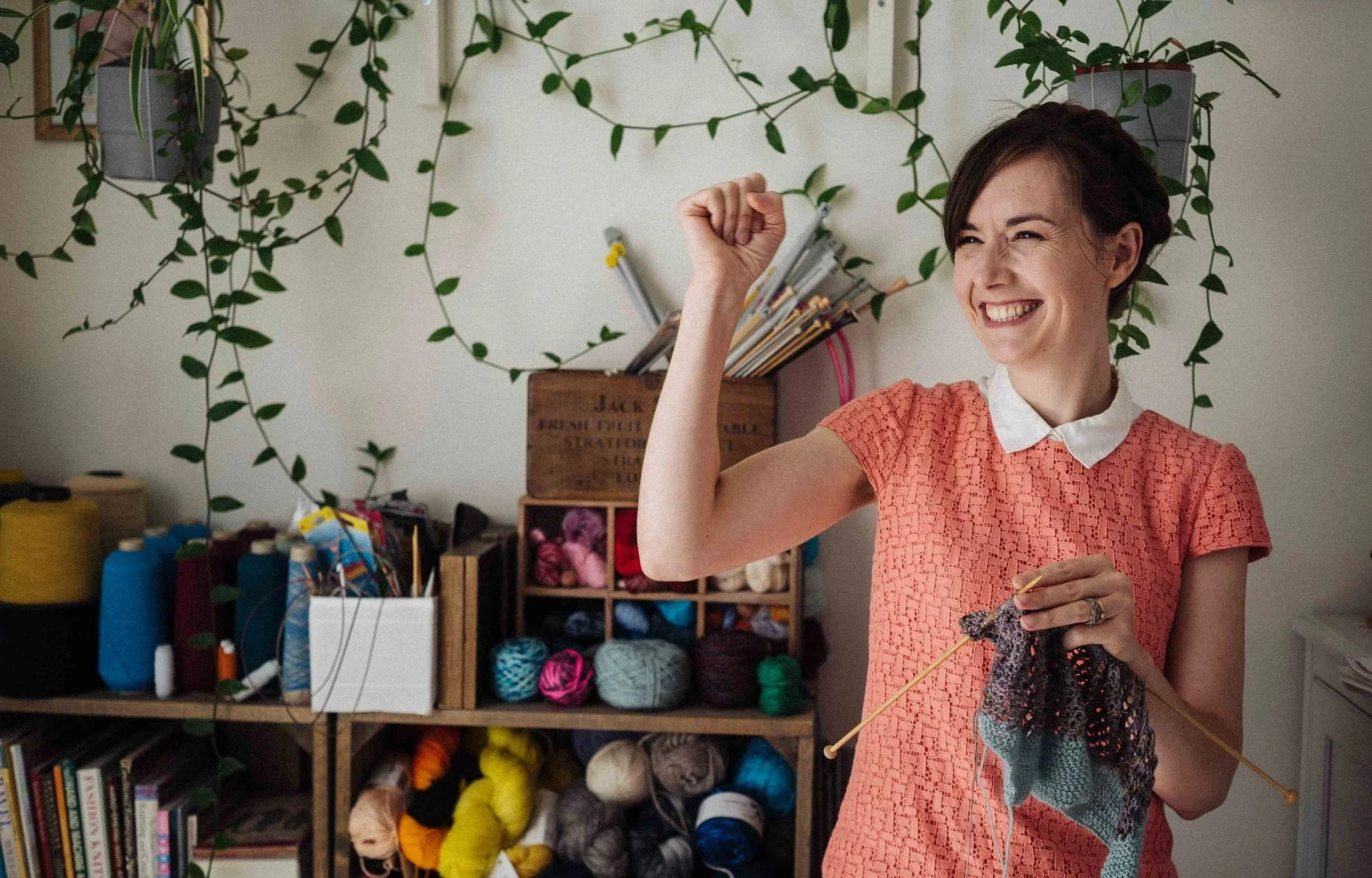 Alice, a white female knitting teacher hold knitting and an arm up triumphantly against a backdrop of yarn and knitting needles