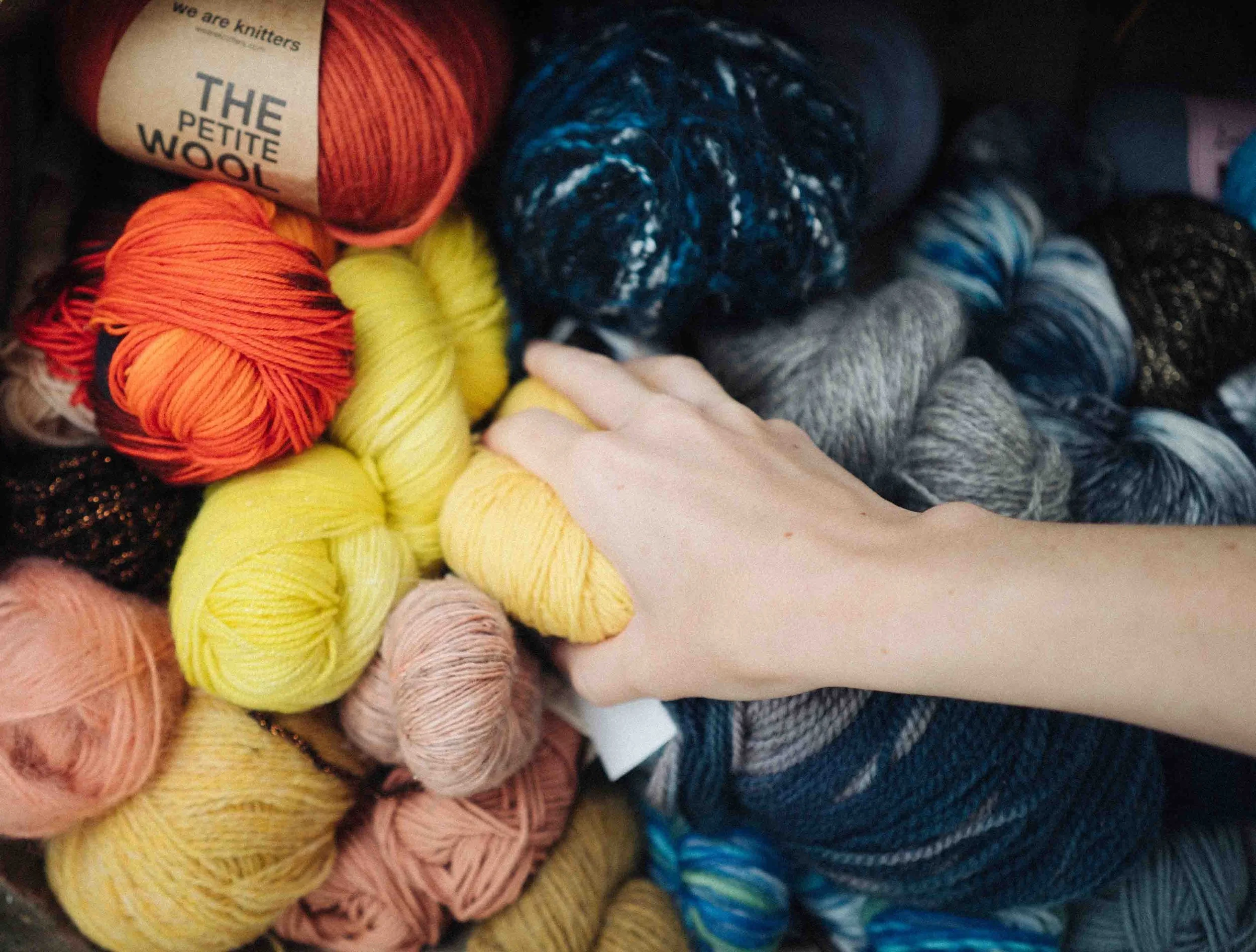 A white female hand reaches into a stack of various coloured knitting yarns
