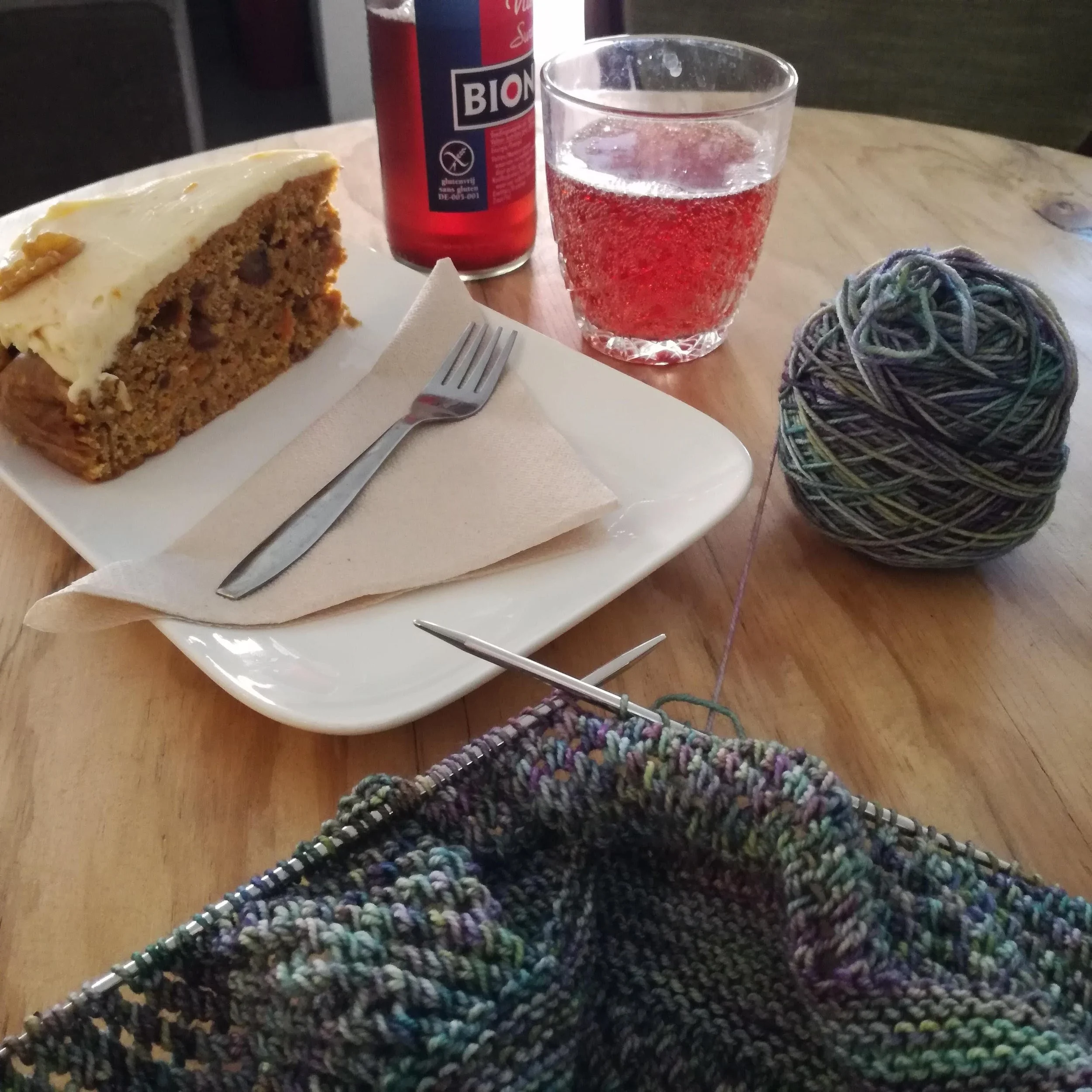 Knitting on metal needles lays on a table next to a piece of cake and a soft drink