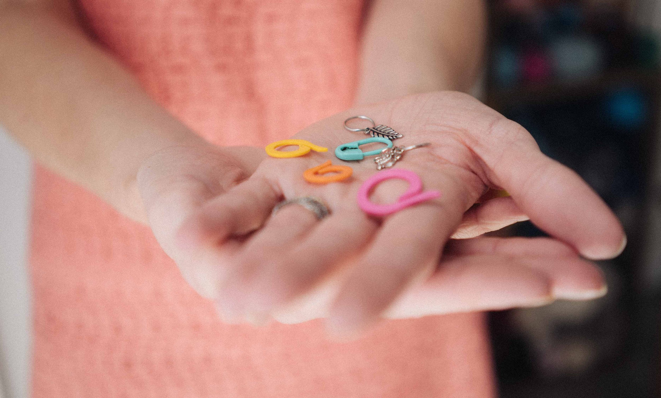 Close-up of a person's hand holding colourful variety of knitting stitch markers against a soft pink background.