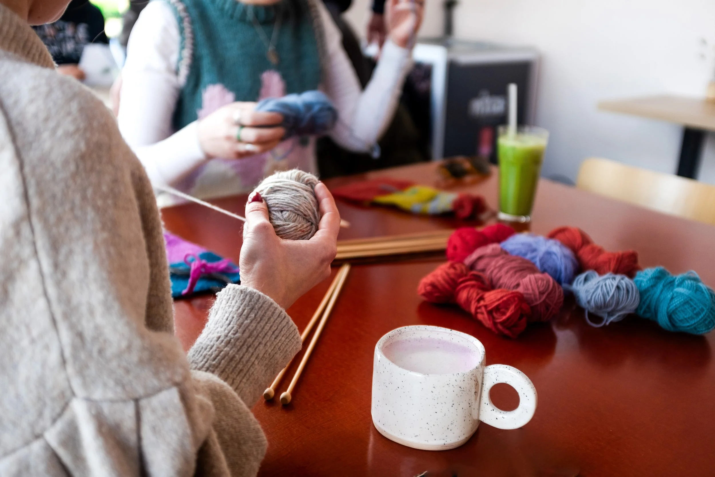Close-up of a person holding a ball of yarn, with other yarn balls and knitting needles on the table, and people in the background knitting or working on yarn projects.