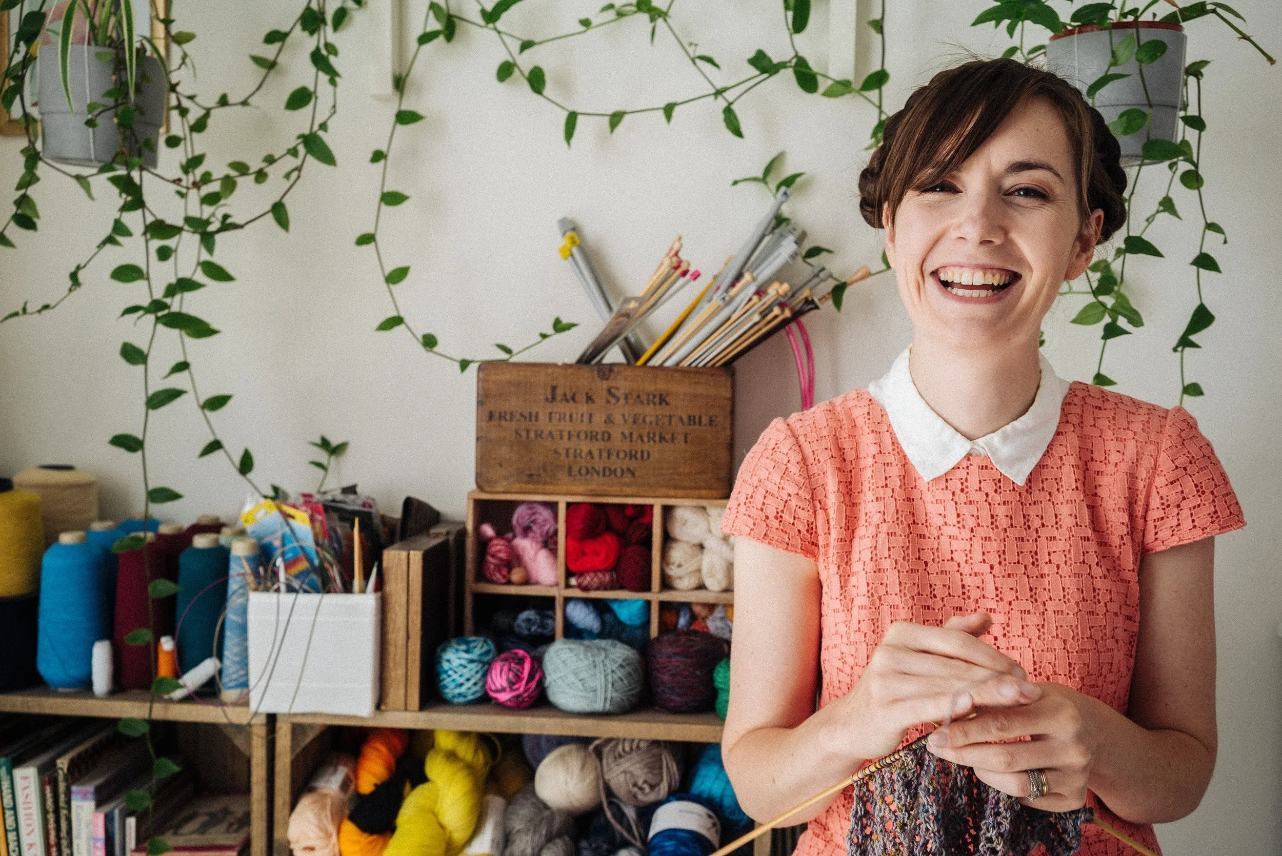 Alice a knitting teacher sits surrounded by yarn and with knitting in her hands, smilling
