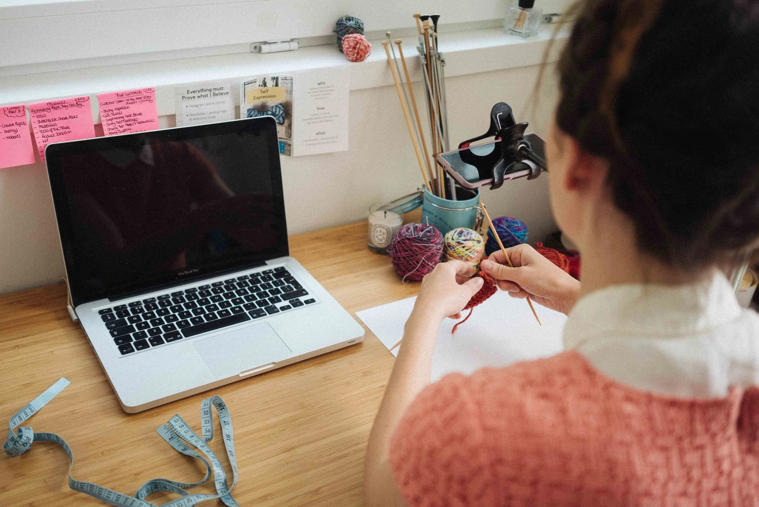 A woman knitting with colorful yarns on a desk next to a silver MacBook laptop, with measuring tapes and knitting supplies around her.