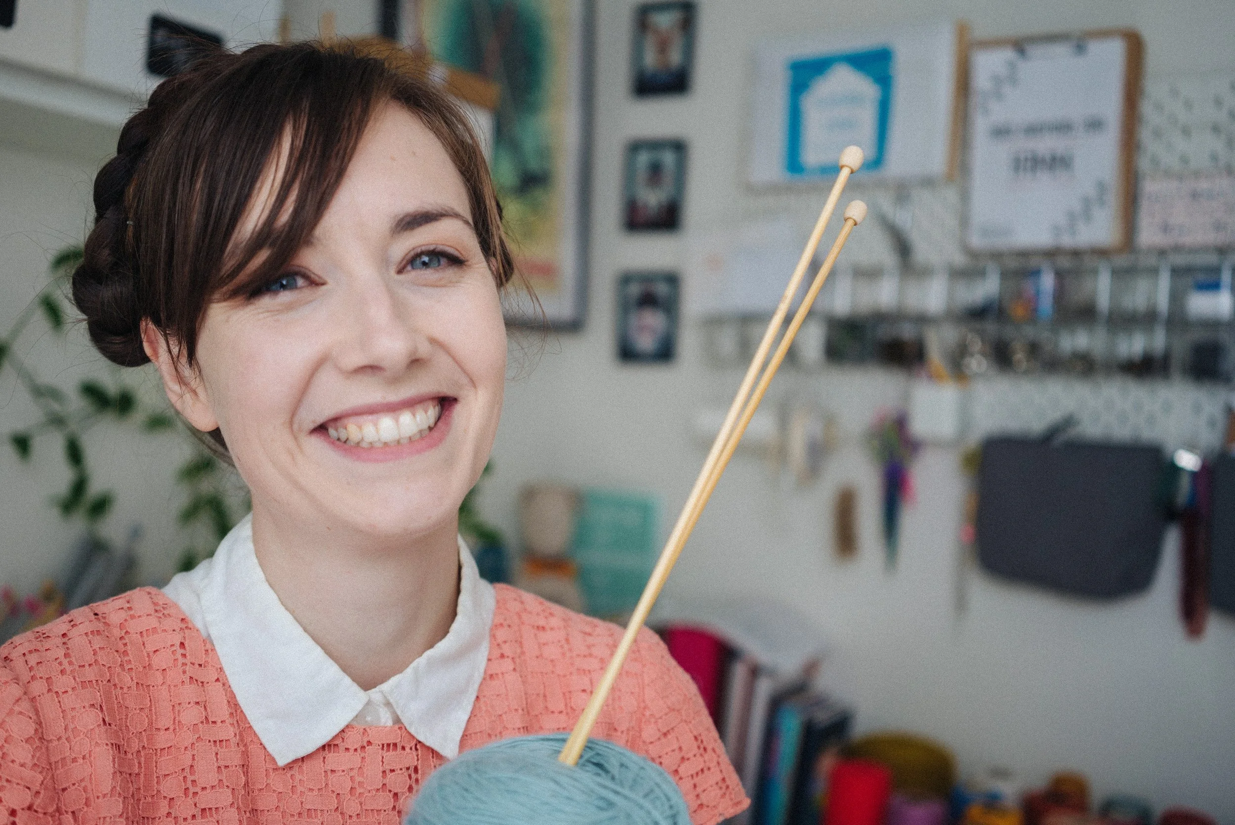 Knitting Teacher Alice smiles while holding blue yarn with wooden knitting needles stuck in it. She wears a peach dress with shite collar