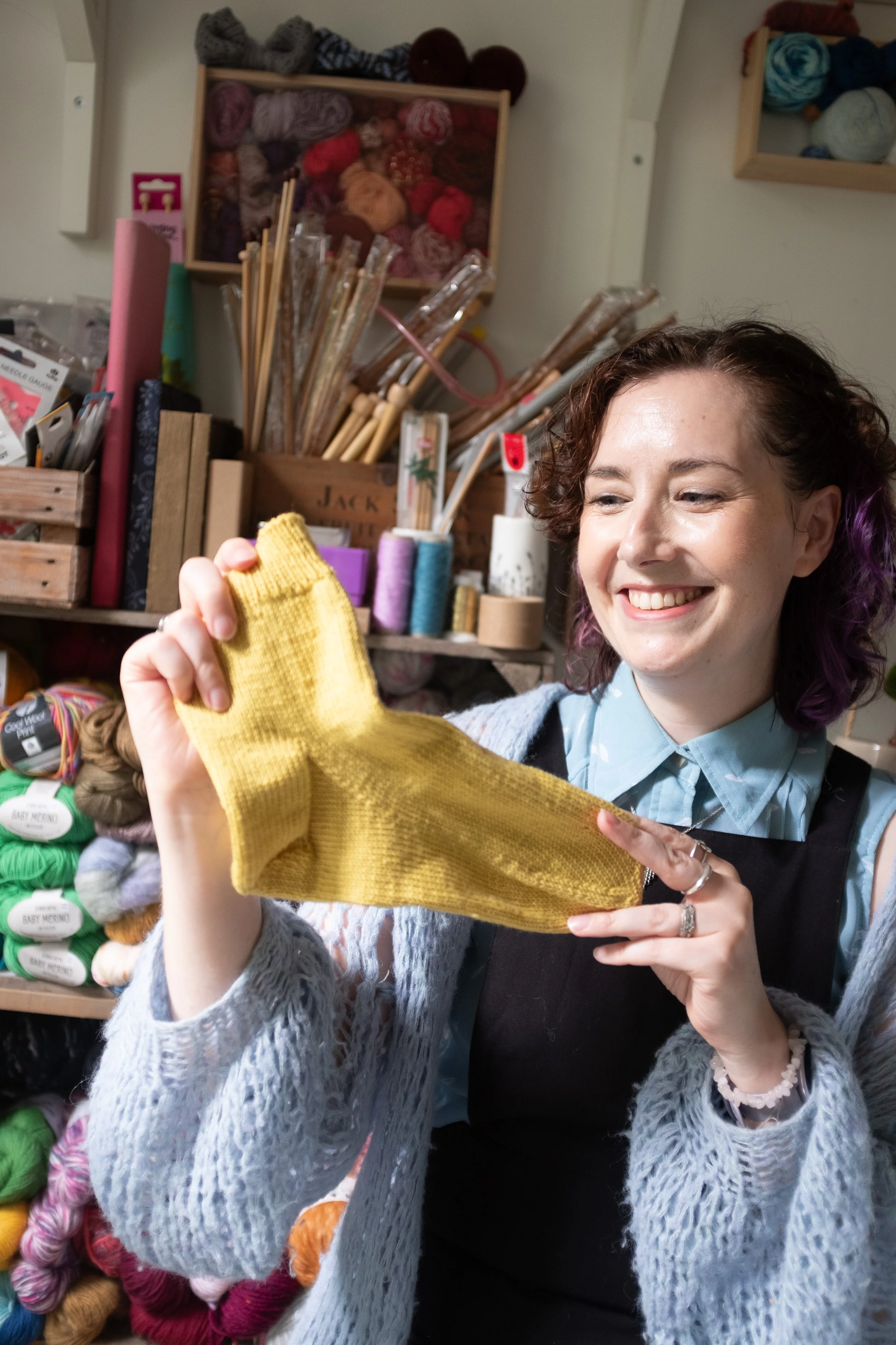 Alice, a knitting teacher holds a yellow hand knitted sock and smiles