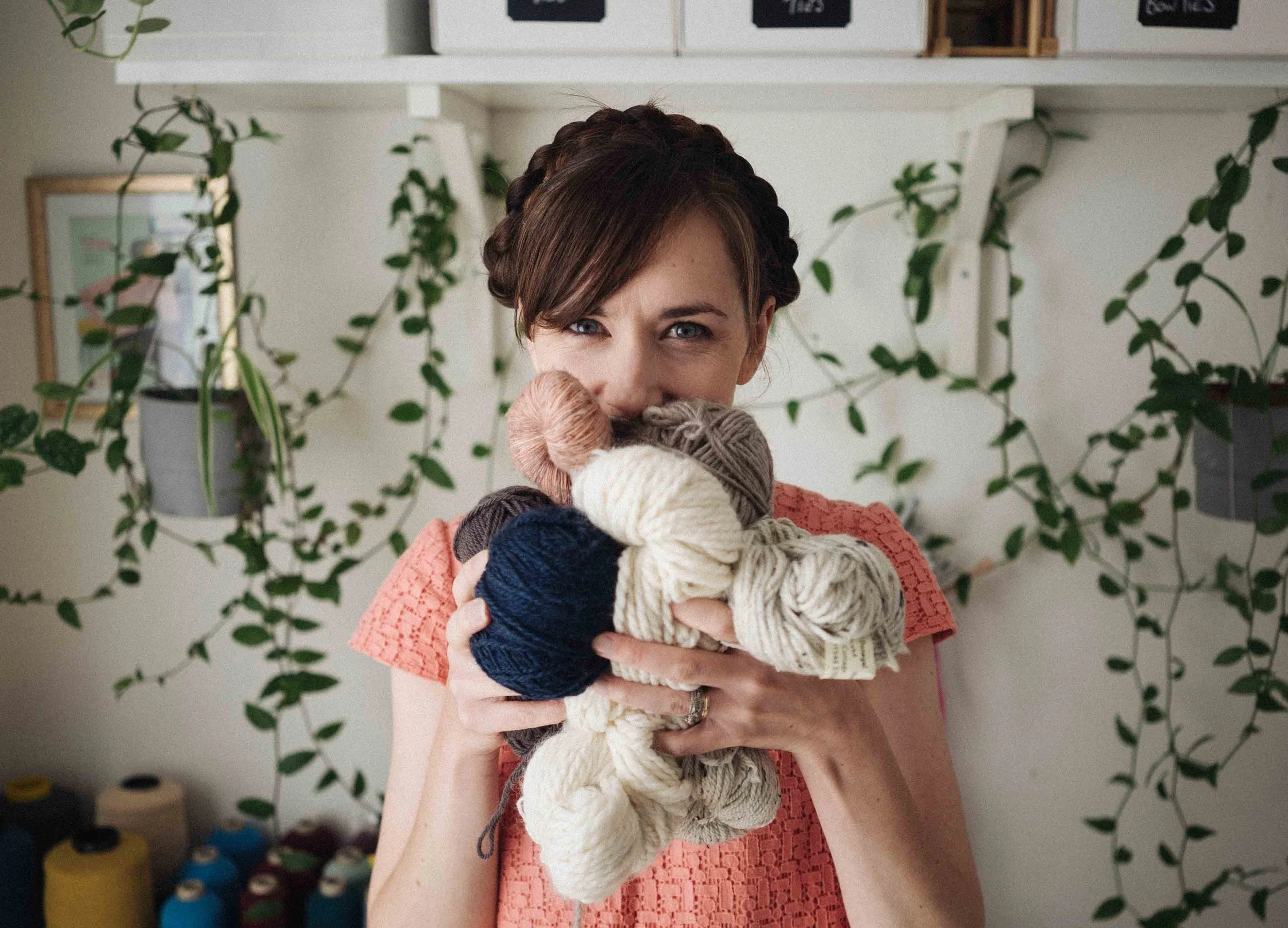 Alice a knitting teacher holds a bundle of knitting yarns covering her mouth. There are vines and yarn cones behind her