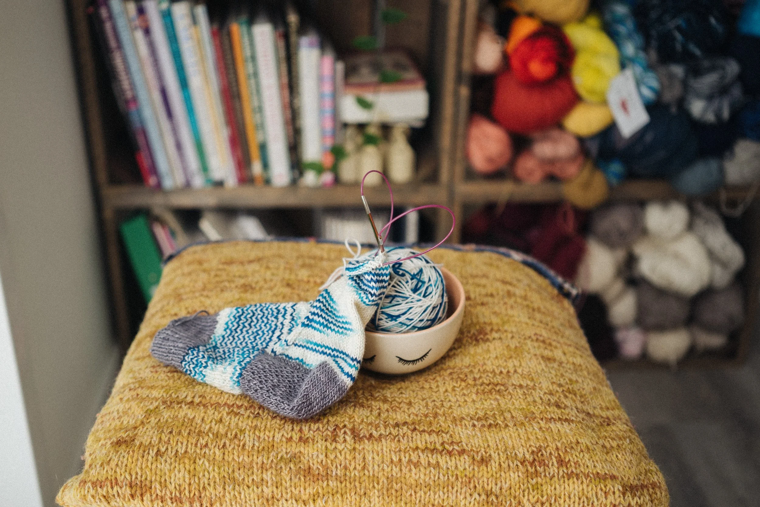 Grey, blue and white hand-knitted sock, partially finished lays on a knitted yellow cushion