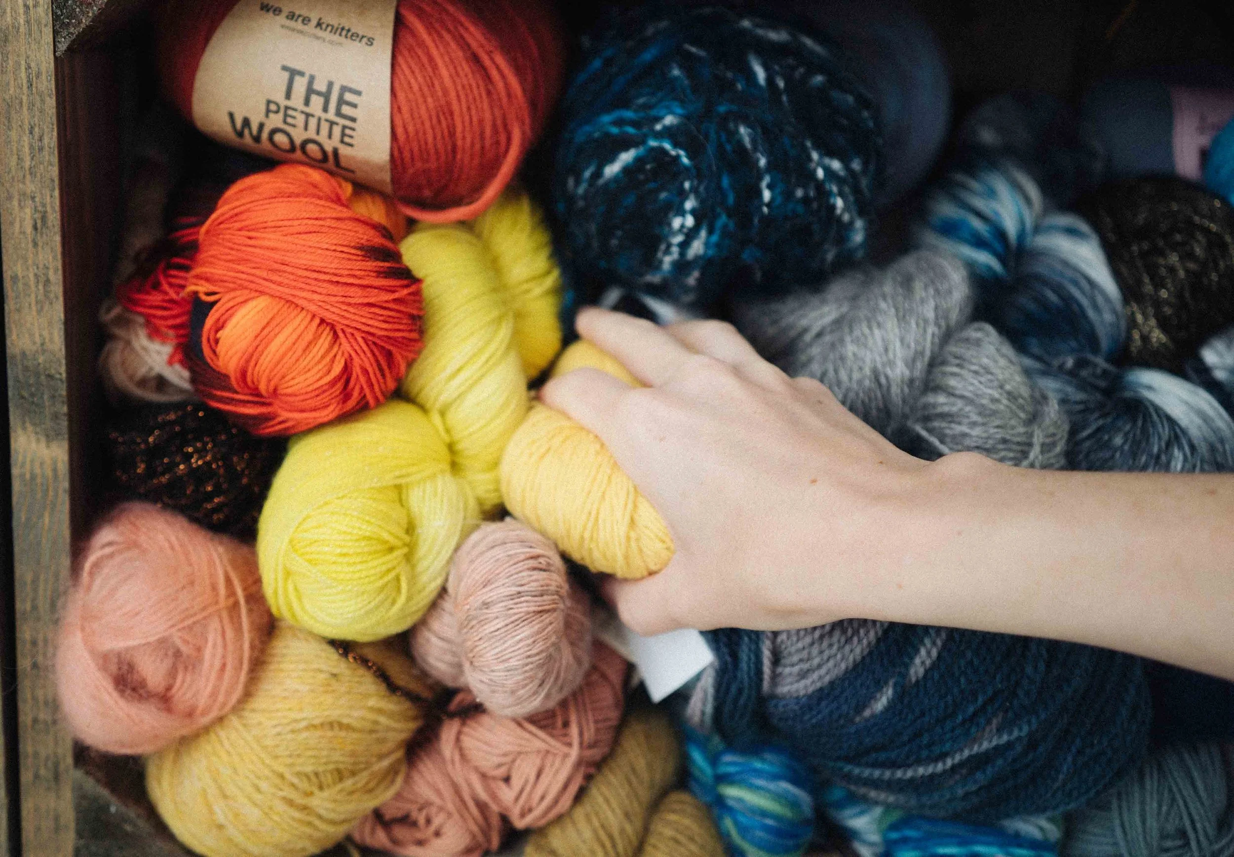 A white female hand reaches into a wooden shelf of yellow and blue and orange yarn.