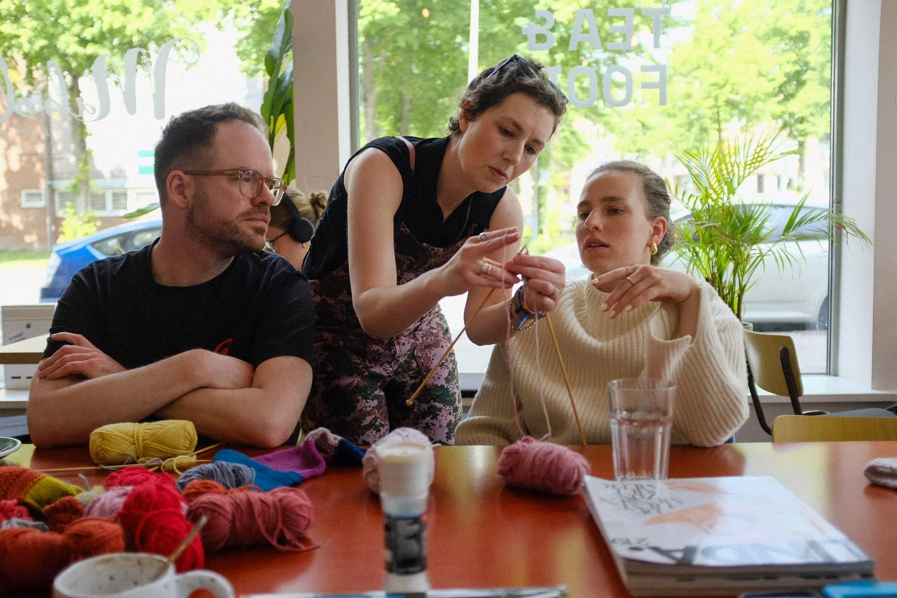 Three people sitting at a table with yarn and knitting supplies, three of the people are focused on yarn and knitting, in a room with large windows and a view of trees outside.