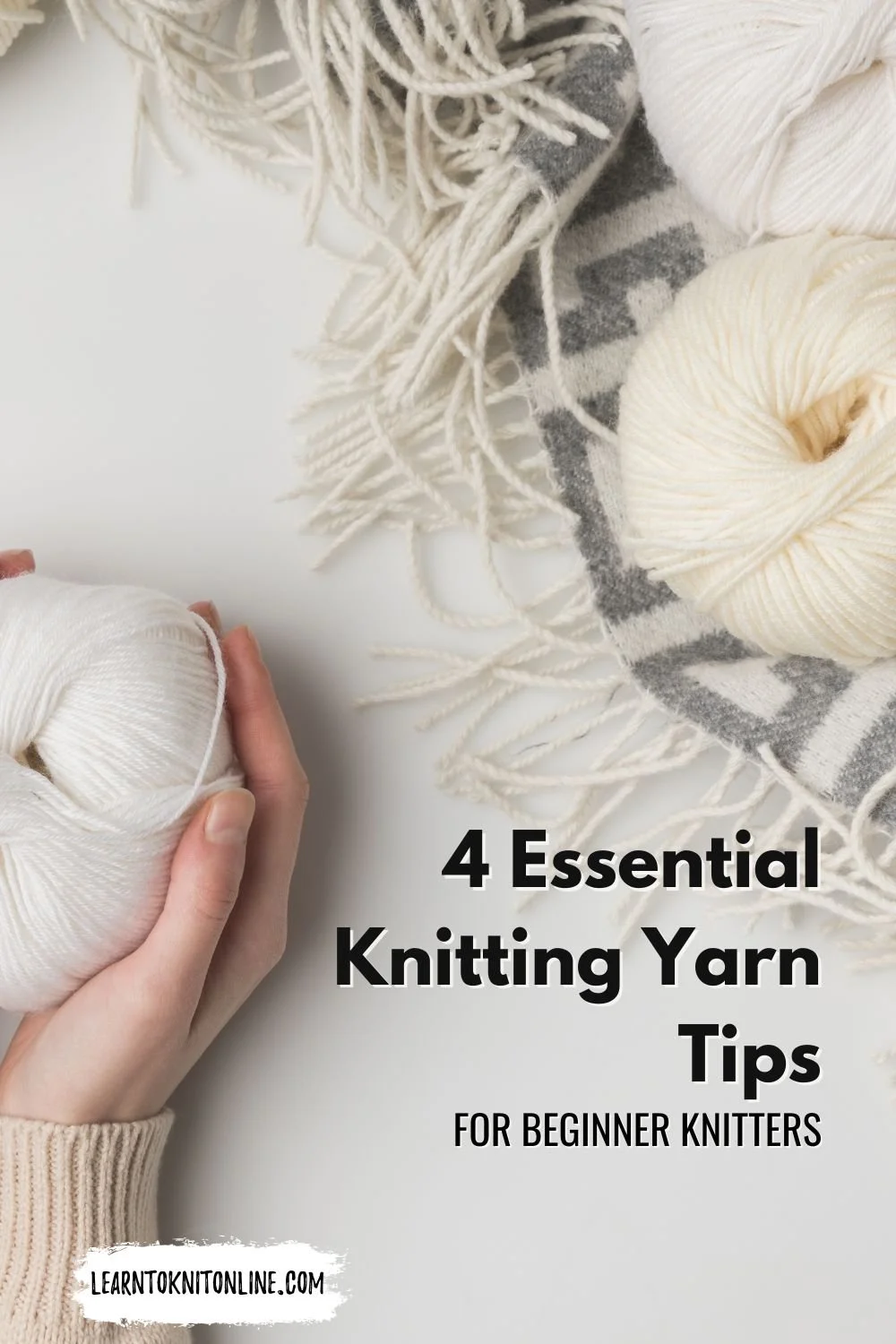 A white hand with a cream sleeve holds a ball of white yarn with cream and white yarn on a grey fringed blanket