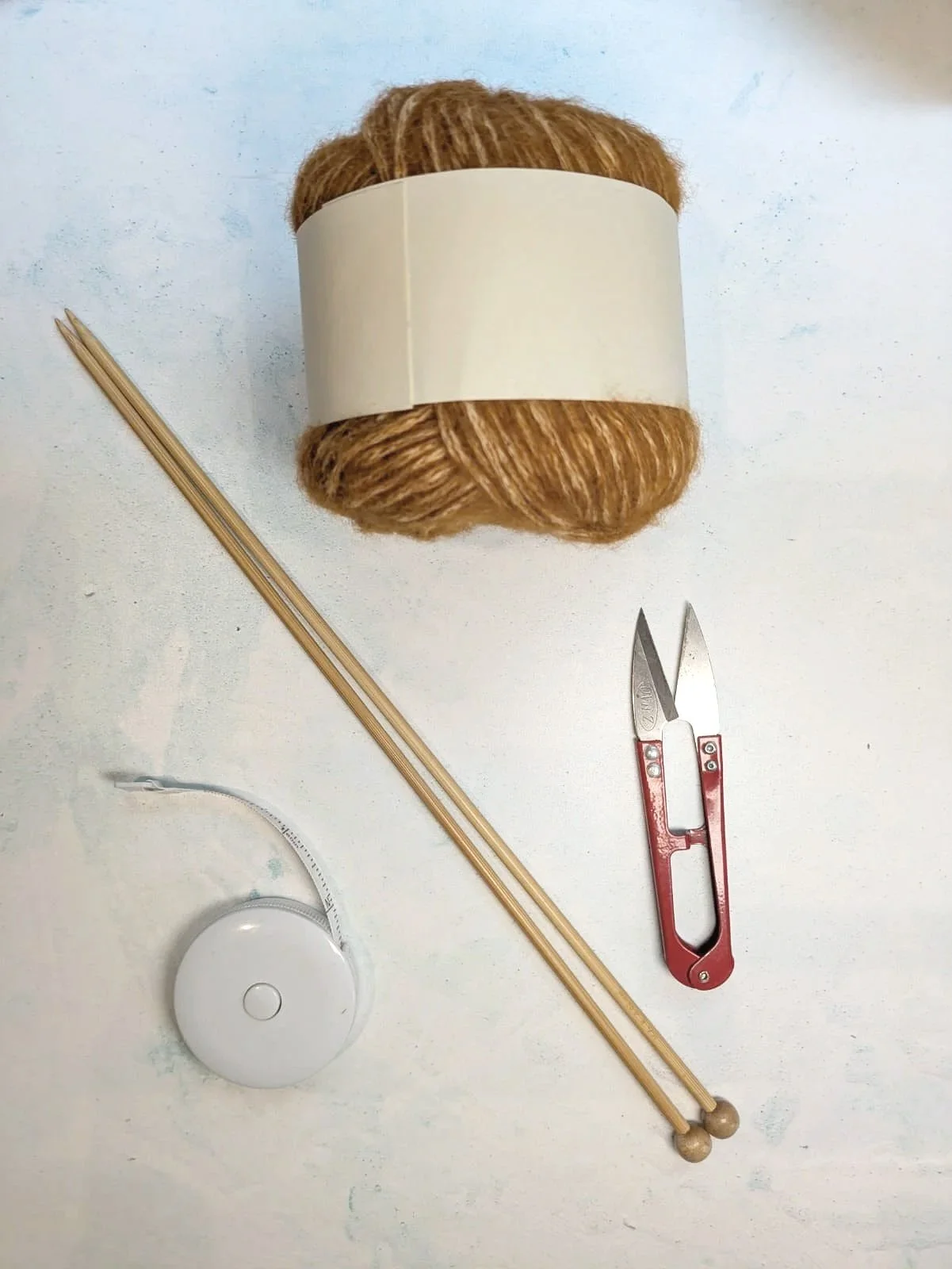 A ball of brown yarn with a white paper band, two wooden skewers, a pair of small scissors, and a white yarn ball winder on a white surface.