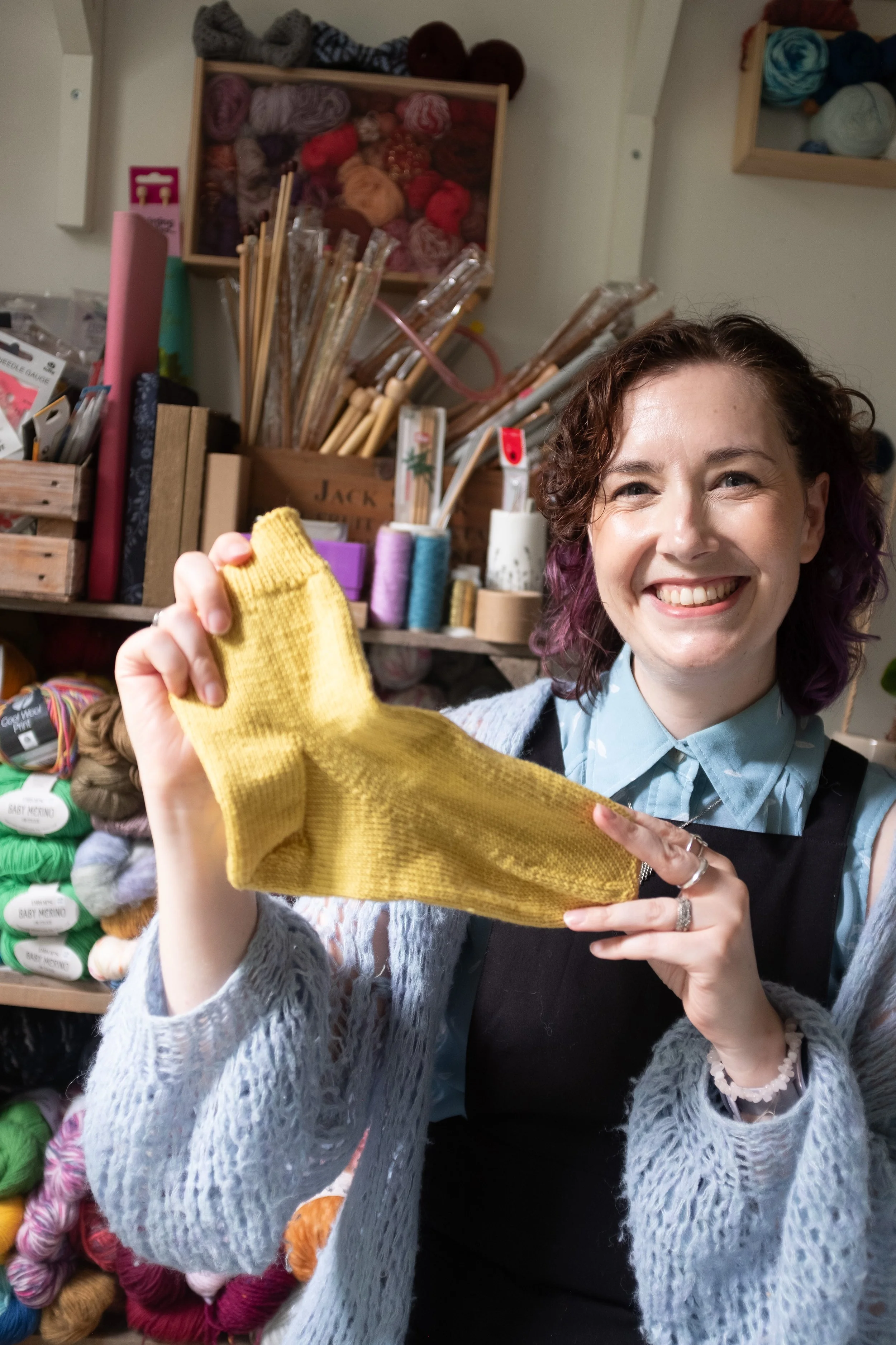 Alice, a knitting teacher holds a yellow, hand-knitted sock and smiles at the camera